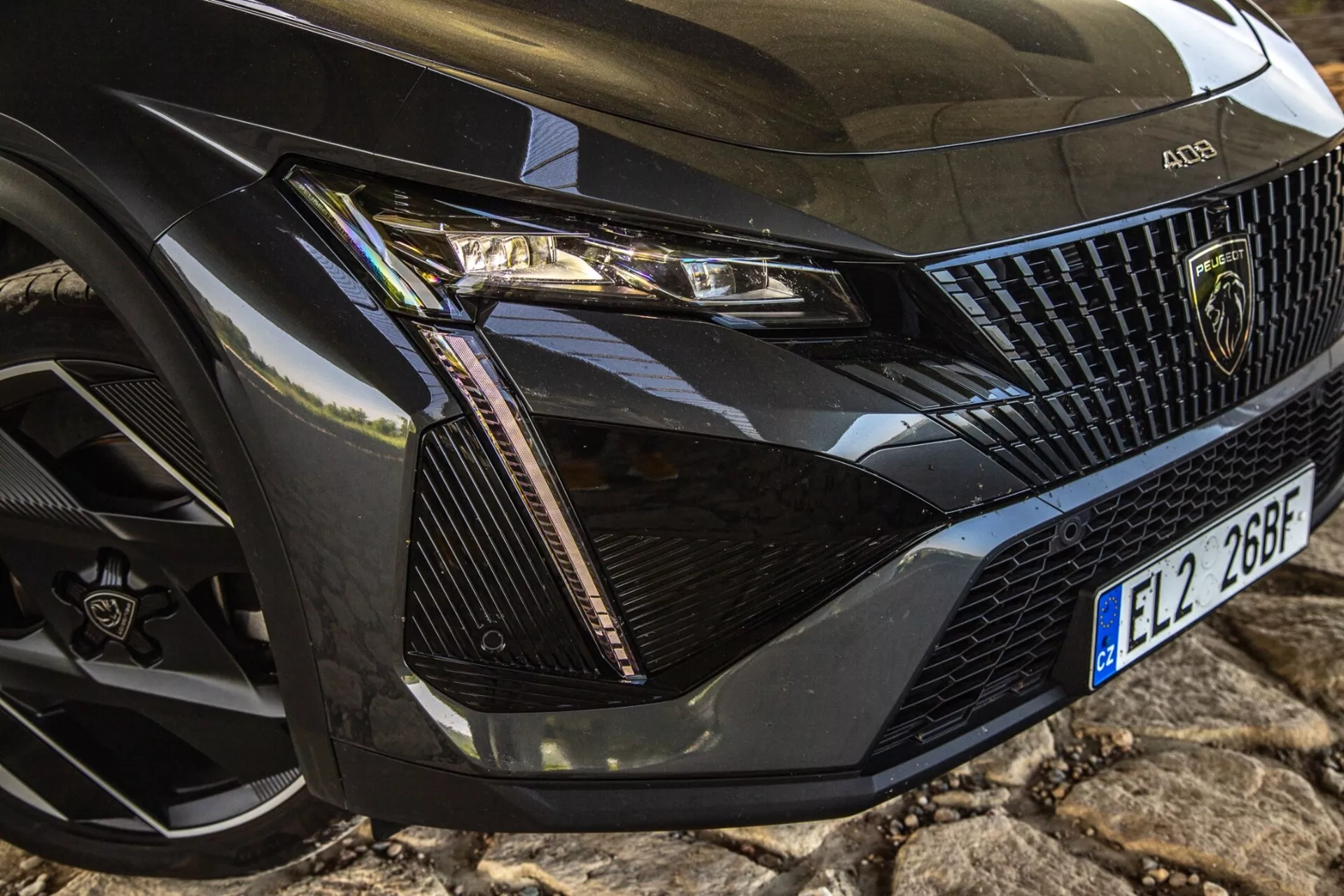 Podroben pogled na sprednji levi kot Peugeota 408 iz leta 2023, ki prikazuje podrobno sestavo žarometa, del rešetke, pokrov motorja in kolo z moderno tehnologijo LED in elementi razkošnega oblikovanja.