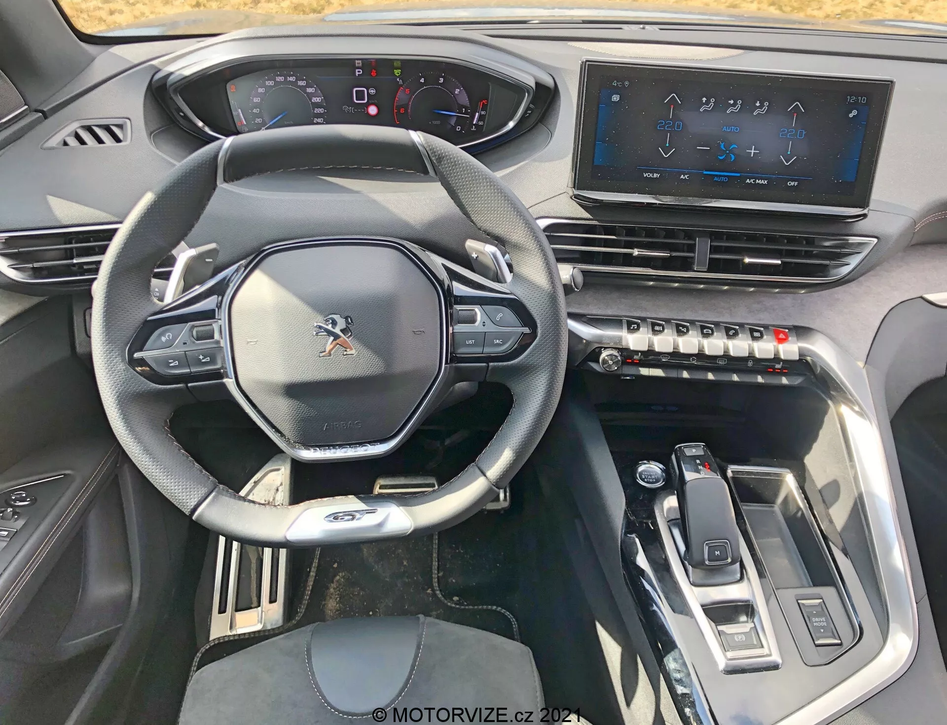 Pogled na notranjost Peugeot 5008 iz leta 2021 s sedeža voznika, ki prikazuje športno večfunkcijsko volan z vgrajenimi krmilniki, delno digitalno instrumentno ploščo, velik zaslon na dotik infotainment sistema, taktilne gumbe za upravljanje klime, eleganten izbirnik prestav in sodobne oblikovne elemente z kovinskimi poudarki za visoko tehnološko, prestižno izkušnjo.