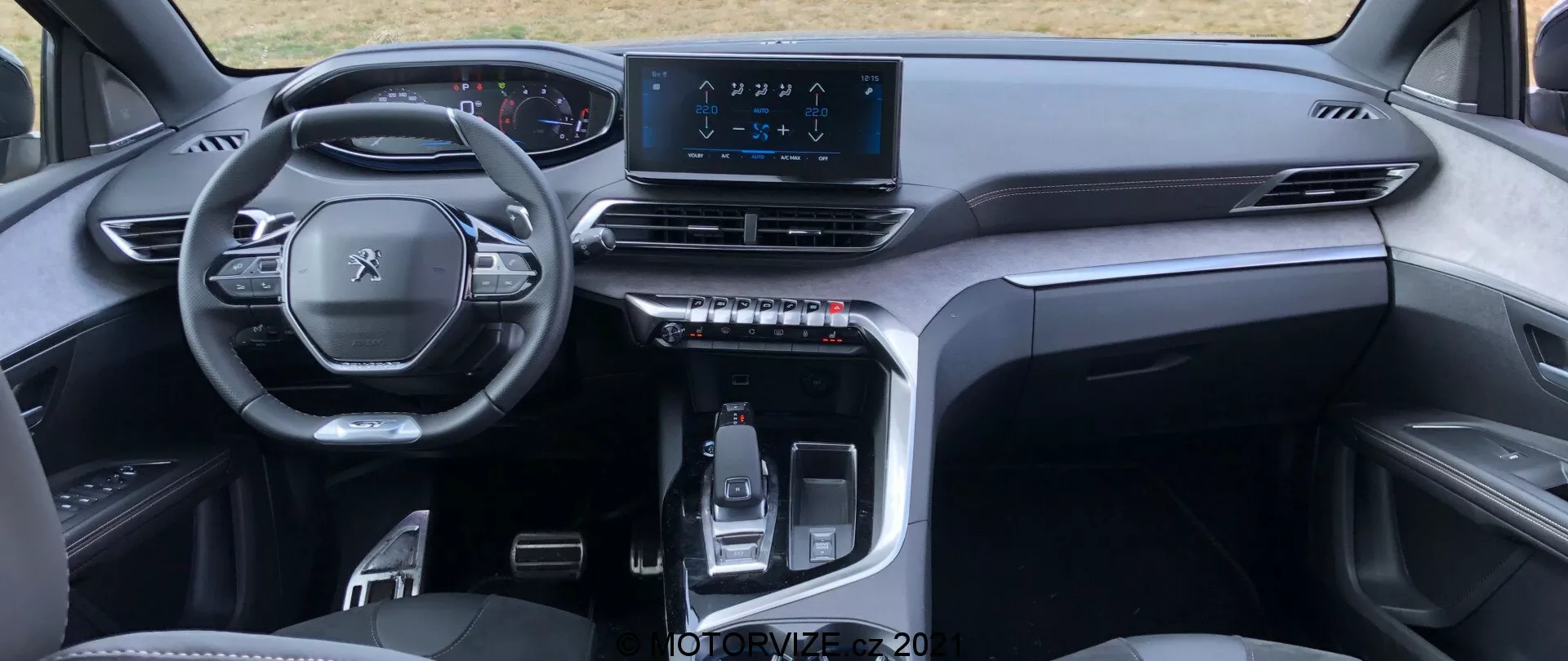 Notranji pogled Peugeot 5008 2021 iz perspektive zadnjega sedeža poudarja večfunkcijski volan, digitalno instrumentno ploščo, simetrično armaturno ploščo z zračniki, osrednji infotainment zaslon na dotik in elegantno sredinsko konzolo z elektronsko krmiljeno prestavno ročico in otipljivimi gumbi. Elegantno minimalistično zasnovo z visokokakovostnimi materiali nakazuje sodobno, premium vozilo.