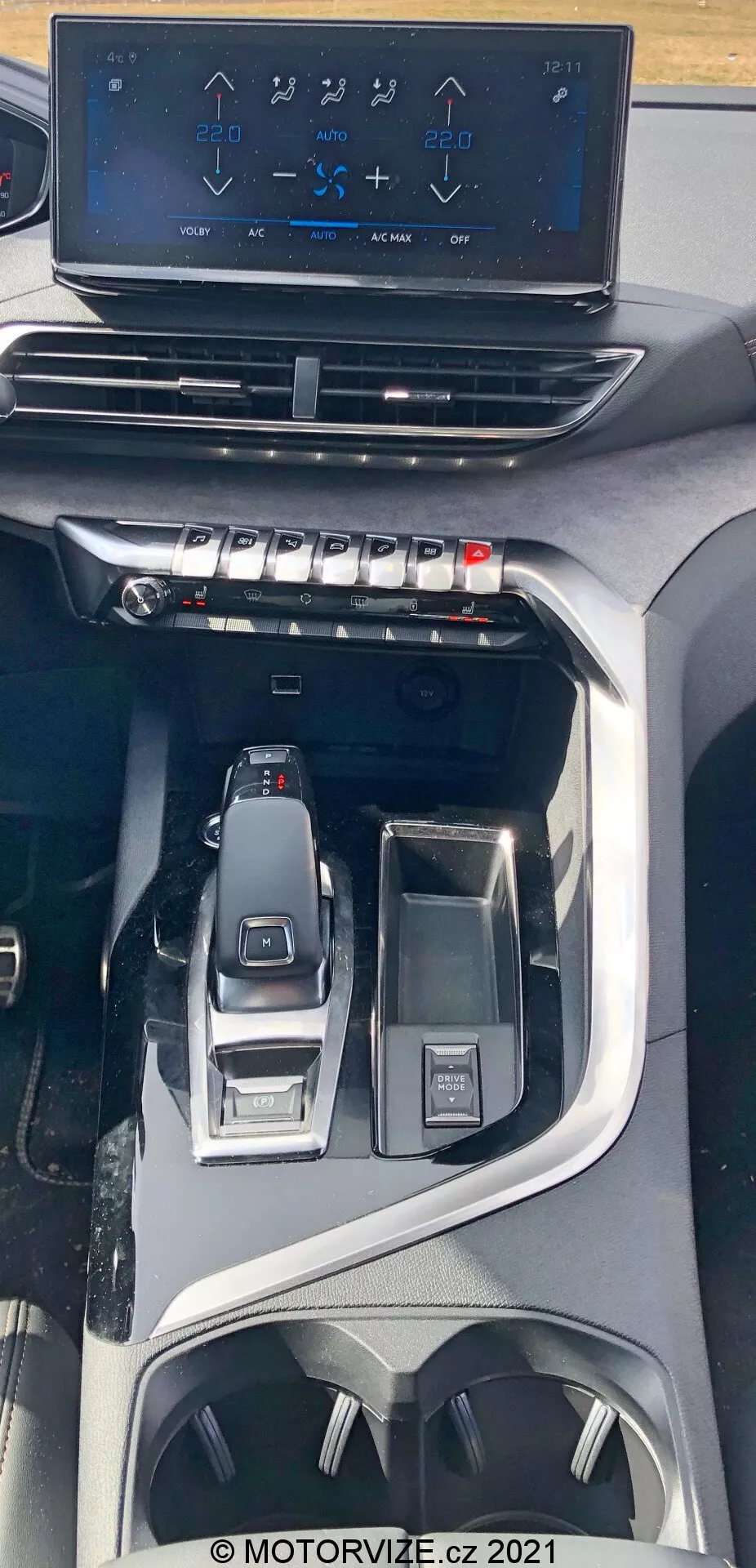 Pogled v notranjost Peugeot 5008 iz leta 2021, ki prikazuje sredinsko konzolo od zgoraj navzdol. Slika poudarja lebdeči info-zabavni zaslon na dotik, ki prikazuje kontrole klimatske naprave nad fizičnimi gumbi in gumbi za hitro nastavitev klimatskih kontrol. Spodaj je videti eleganten elektronski ročni menjalnik z bližnimi kontrolami za izbiro voznega načina in večfunkcijsko nadzorno tipko. Dizajn nakazuje sodoben, premium avto z naprednimi tehnološkimi in ergonomskimi funkcijami, vključno s držali za pijače in shranjevalnimi prostori.