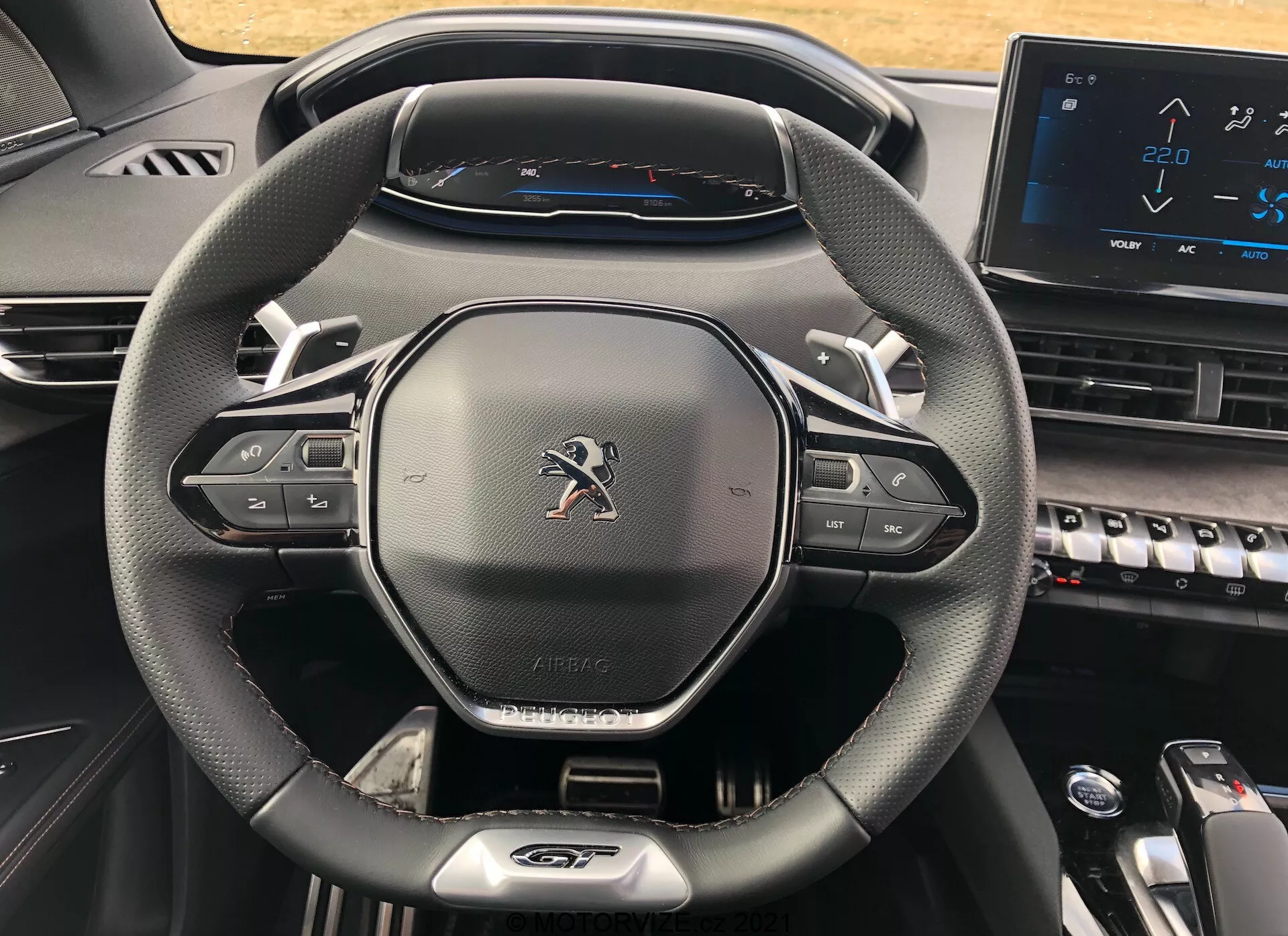 Pogled od blizu na voznikov kokpit v modelu Peugeot 5008 iz leta 2021, s poudarkom na sodobnem trikrake volanu z usnjeno teksturo, gumbi za upravljanje z multimedijo in tempomatom, Peugeotovim emblemom, oznako 'AIRBAG', popolnoma digitalni instrumentni plošči, ki prikazuje merilnik hitrosti z največjo hitrostjo 220 km/h, in del sredinske konzole s klavirskimi tipkami in elektronskim zaslonom na dotik, kar predstavlja sodoben in tehnološko napreden dizajn.