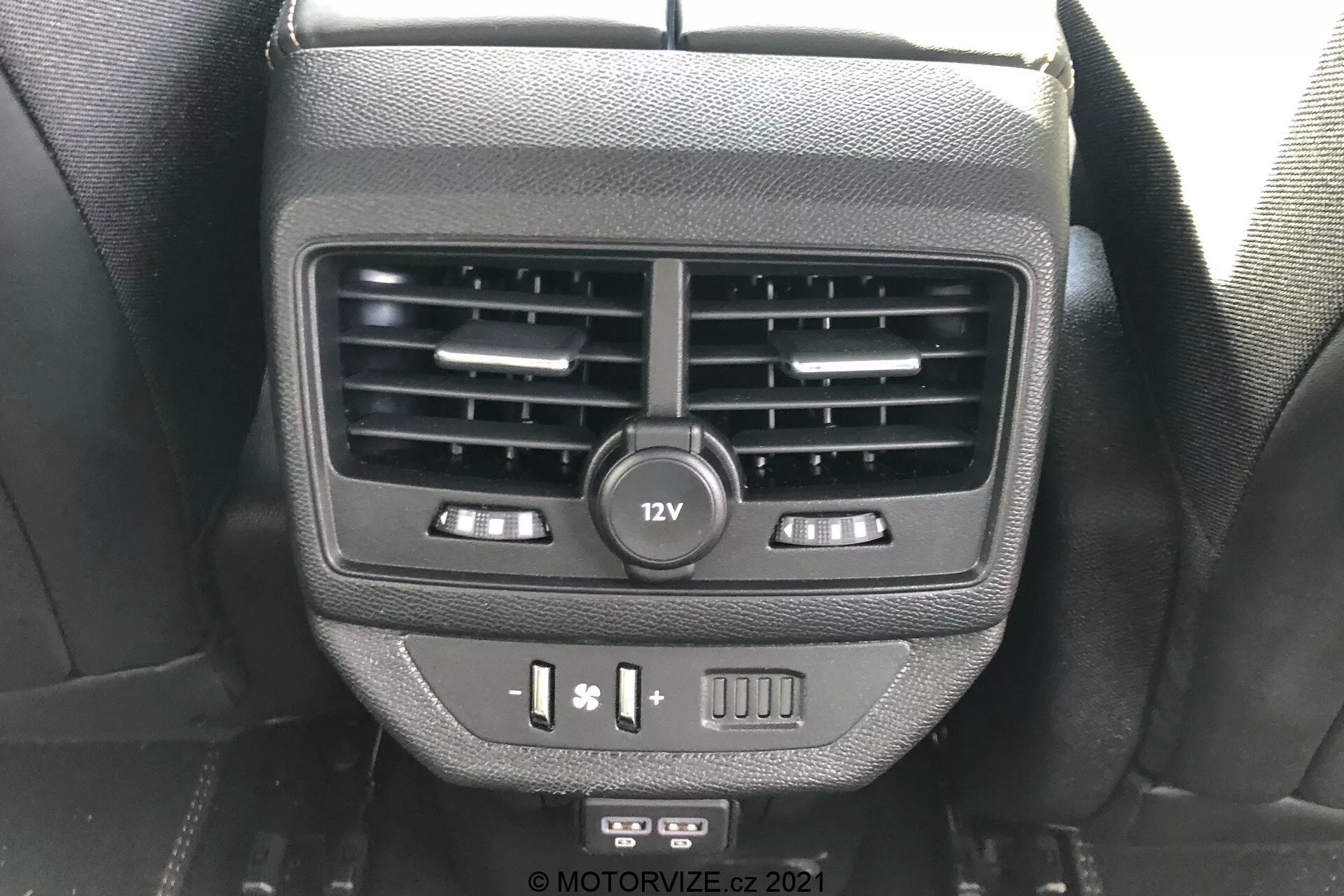 Podroben pogled na funkcije za zadnje potnike na zadnji strani sredinske konzole v Peugeot 5008 2021. Slika prikazuje dvojna prezračevalna kanala, 12V vtičnico, kontrole za ogrevanje sedežev, USB priključke in prazno tipko, posneta z vidika zadnjega potnika.