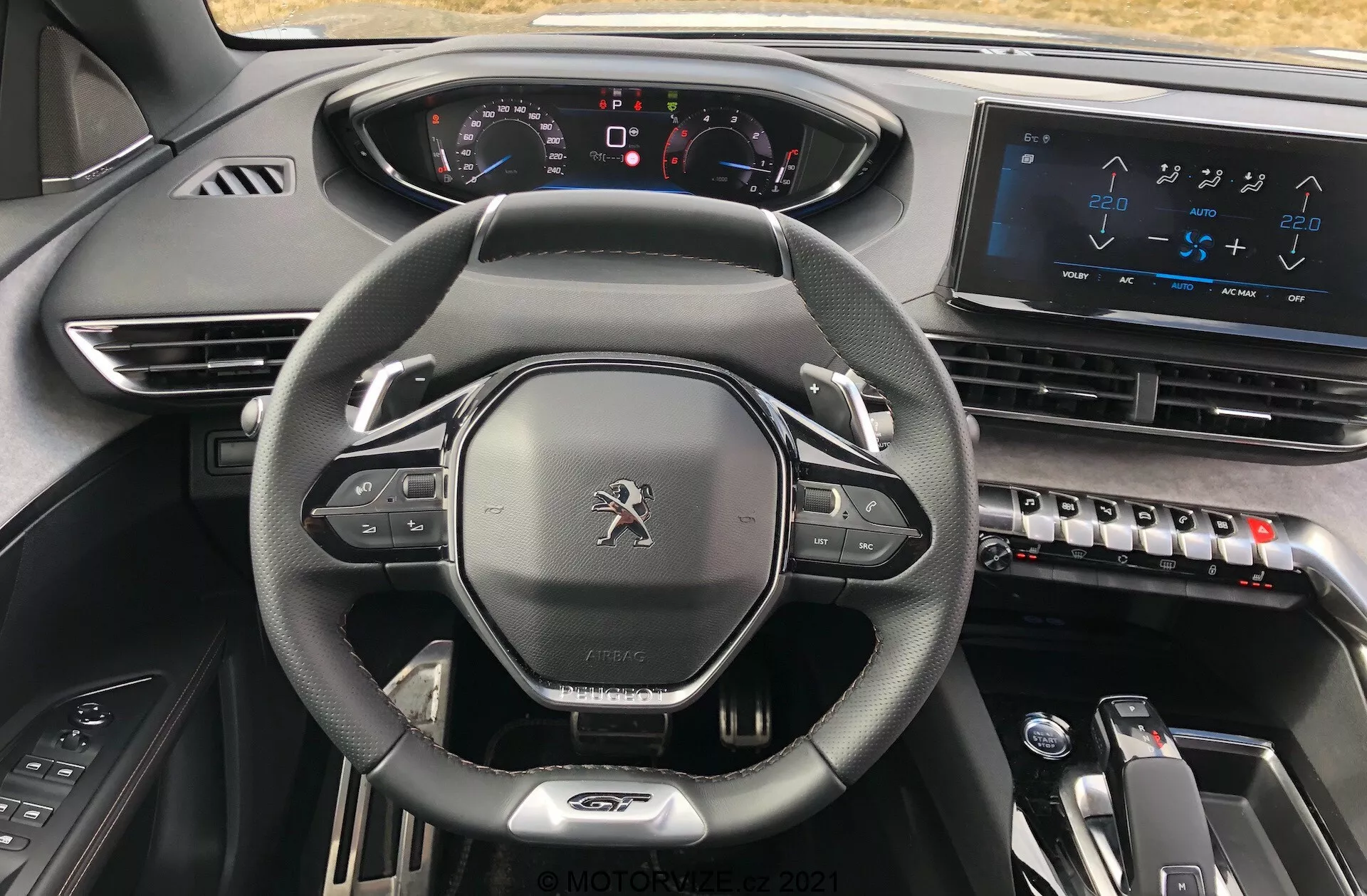 Notranji pogled Peugeot 5008 2021 iz sedeža voznika, ki poudarja usnjen volan s ploskim dnom z vgrajenimi kontrolami, popolnoma digitalno armaturno ploščo z osvetljenimi kazalci, velik zaslon na dotik z informacijskim in zabavnim sistemom na sredinski konzoli, elegantne kontrole klimatske naprave, elektronski izbirnik prestav za samodejni menjalnik in premium oblikovno zasnovo temnih materialov, ki izraža moderno, napredno in luksuzno estetiko.