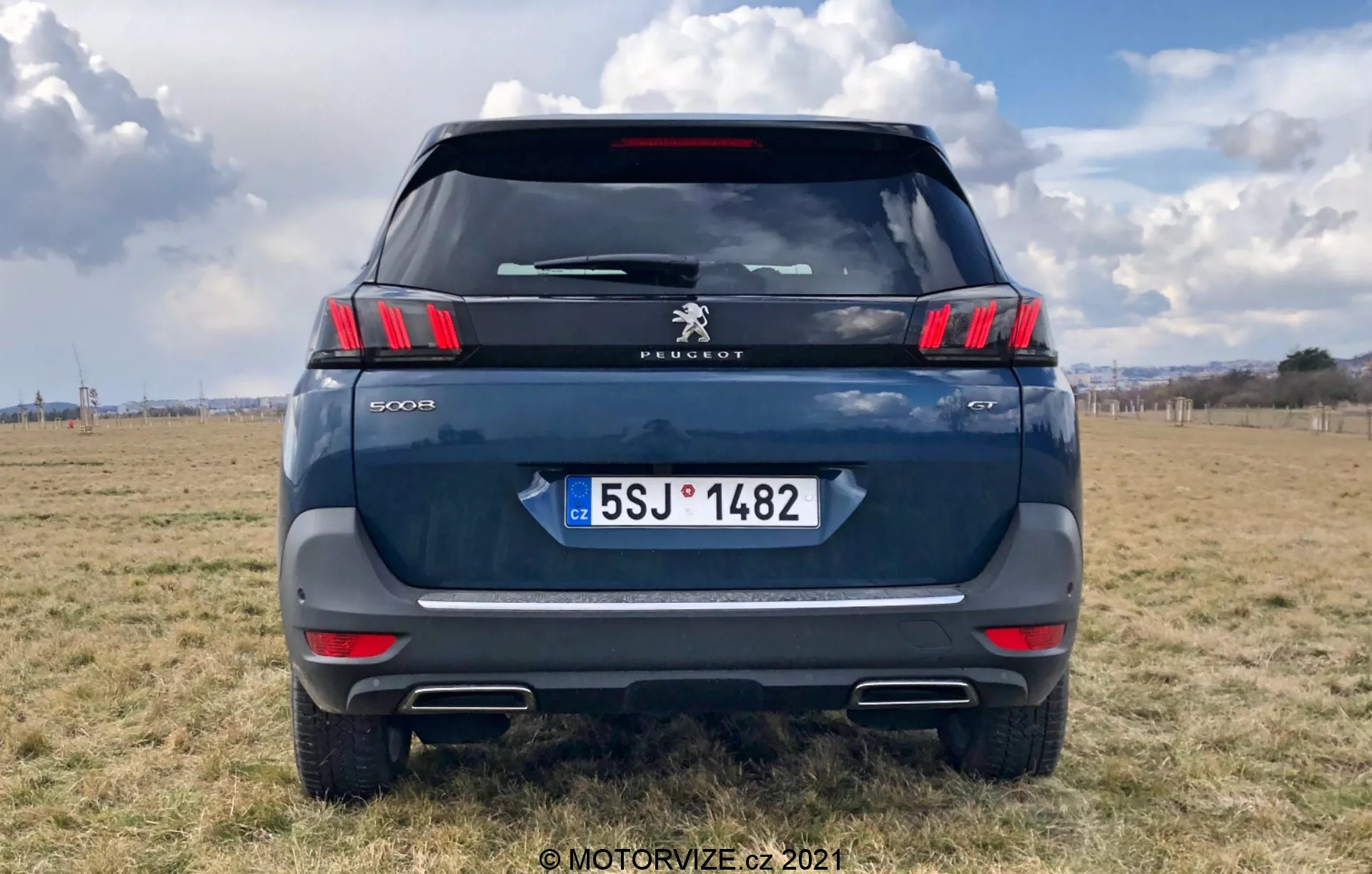 Zadnji pogled na sodoben SUV Peugeot 5008, ki poudarja elegantne horizontalne zadnje luči z značilnimi LED segmenti v obliki krempljev, logotip Peugeot in oznako modela '5008'. Temno modra ali siva barva, parkirano na terenu na prostem s delno oblačnim nebom. Vidni elementi vključujejo zadnje vetrobransko steklo, zadnji brisalec, visoko nameščeno zavorno luč, kljuko prtljažnika in registrsko tablico Češke republike z 'CZ'. Odsevni trak nad odbijačem s parkirnimi senzorji in izrezi izpušnih konic so očitni. Posnetek je osredotočen s avtomobilom, ki zapolni večji del okvirja.