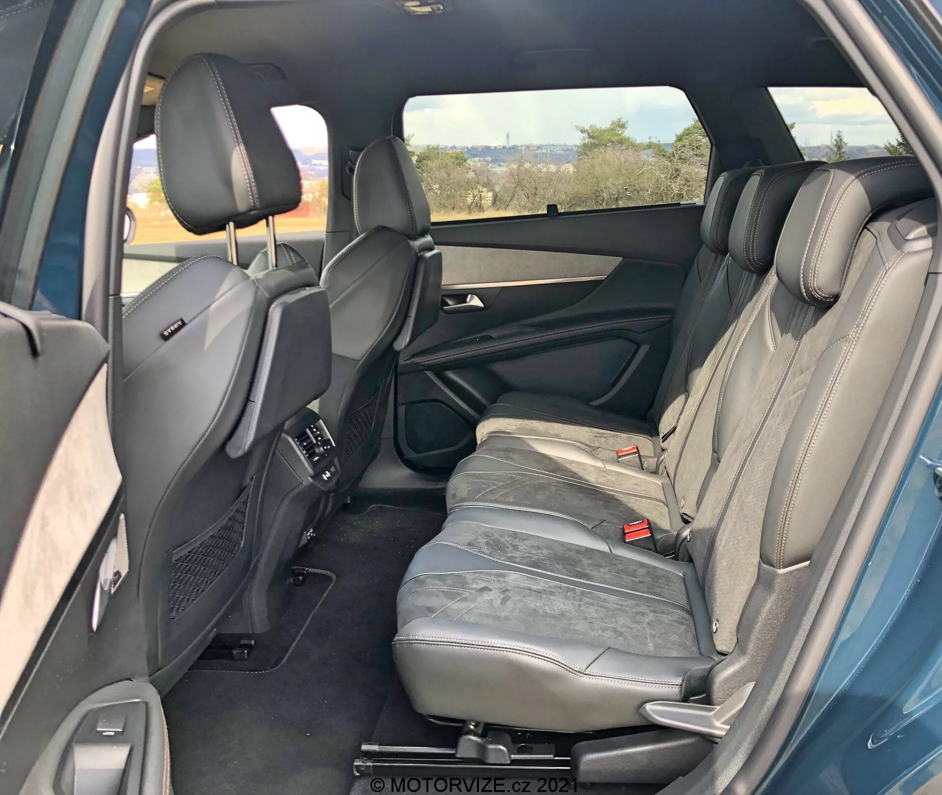 Notranjost Peugeot 5008 2021 iz desnega zadnjega kota, ki prikazuje zadnji potniški prostor. Desna zadnja vrata so odprta, kar razkriva značilnosti vratne obloge, kot so ročaj, shranjevalni predali in zvočnik. Vidni so trije zadnji sedeži, oblazinjeni s sivo usnje, z naslonjali za glavo. Osrednji sedež naslon za roke je v pokončnem položaju. Prav tako so vidni zadnji varnostni pasovi in ​​prednji sedeži z nastavljivimi naslonjali za glavo in osrednjo konzolo. Zadnje klimatske odprtine na sredinski konzoli, zatemnjeno desno okno, pogledi na leva zadnja vrata, ki nakazujejo simetrijo. Skupno dobro vzdrževana notranjost brez vidnih tehnoloških lastnosti.