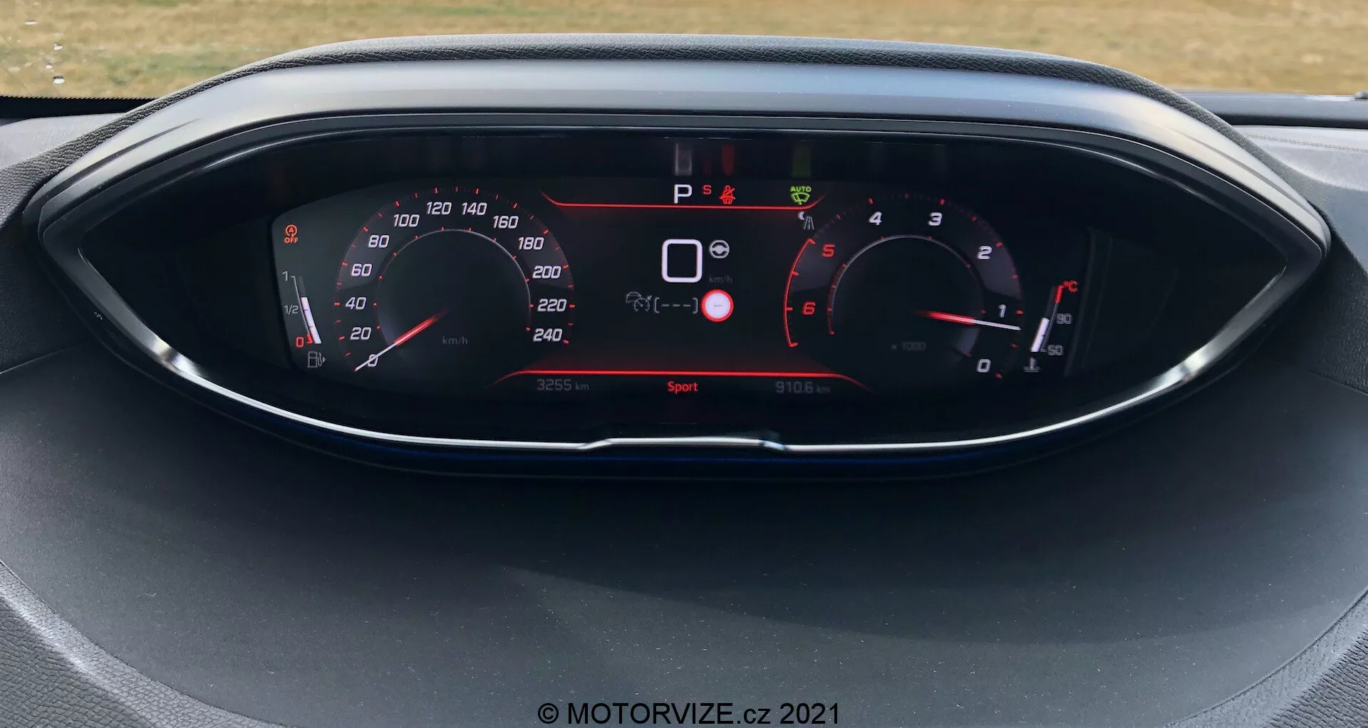Pogled od blizu na instrumentno ploščo Peugeot 5008 iz leta 2021, z digitalnim prikazovalnikom in analognimi kazalniki. Prikazovalnik prikazuje merilnik hitrosti, merilnik vrtljajev, izbor prestave in različne kazalnike vozila, kot so varnostna opozorila in način vožnje. Avto je v mirovanju z aktivnim načinom vožnje 'Sport'.