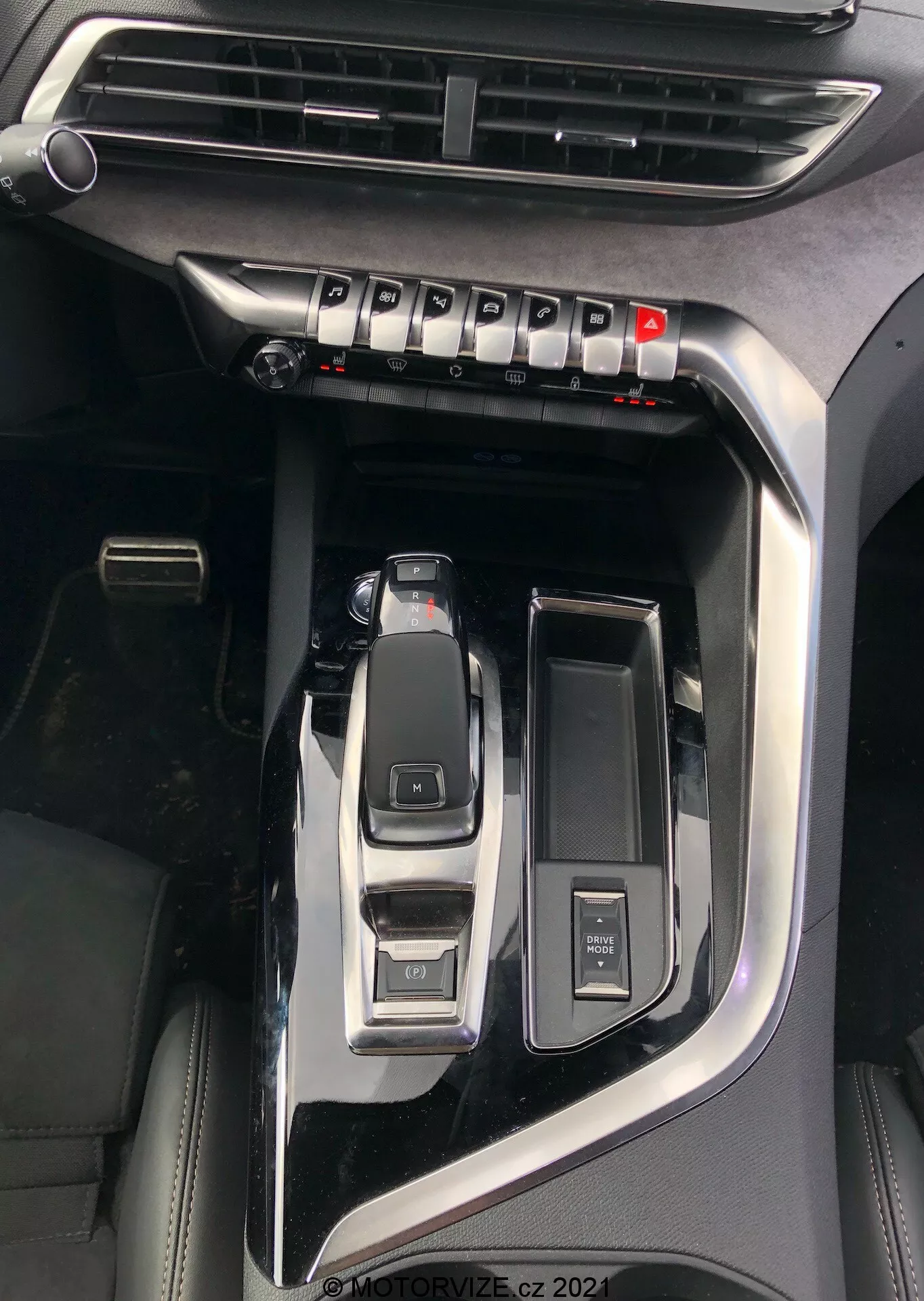 Pogled od blizu na sredinsko konzolo Peugeot 5008 2021, ki poudarja ključne lastnosti, kot so izstopajoča avtomatska prestavna ročica v parkirnem položaju, izbirnik načinov vožnje za načine eco, sport, comfort, gumb za zagon/ustavitev motorja za zagon brez ključa, večfunkcijski krmilni gumb, odprt držalo za skodelico, vidno zračilo za nadzor klime in piano črna obdelava z kovinskimi poudarki, kar kaže na premijsko različico vozila, vodni žig 'MOTORVIZE.cz 2022'.