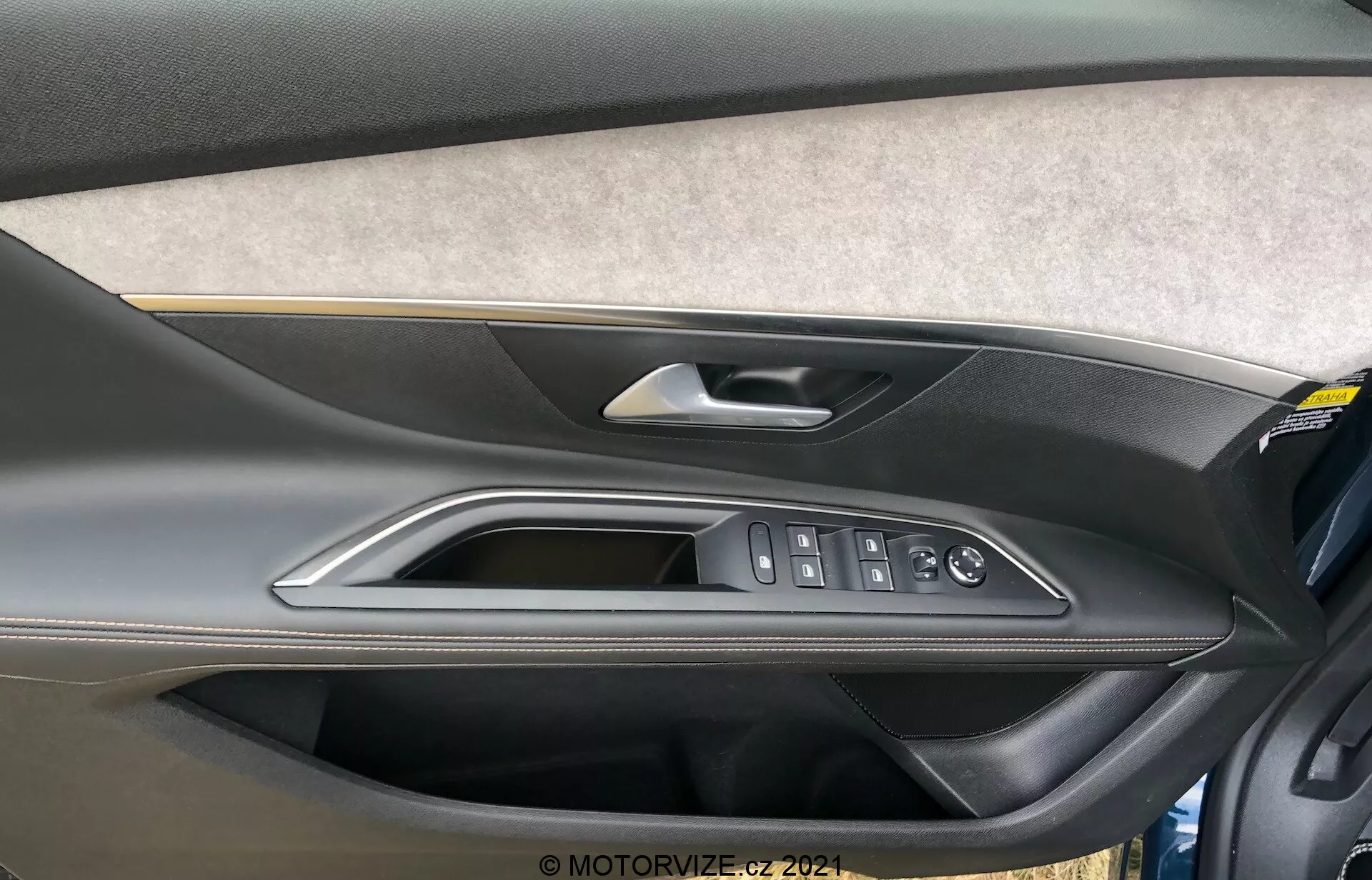 Pogled od blizu na notranjo ploščo vrat Peugeot 5008 2021, ki vključuje odsek oblazinjen s filcom ali tkanino, dvobarvno obliko iz oblikovane plastike s temnejšim zgornjim delom in svetlejšim spodnjim delom, visokokakovostno kovinsko ali kromirano kljuko vrat, polirane obrobe in ergonomske kontrole za električna okna, zaklepanje vrat in stranska ogledala. Vidno je tudi shranjevalni žep na spodnjem delu vratne plošče.