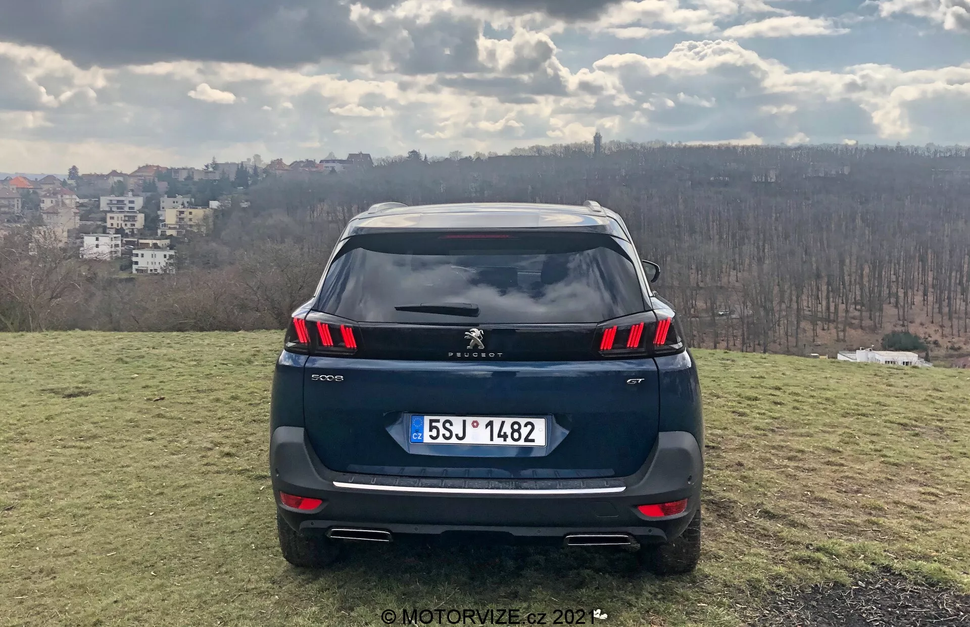 Razen zadnji pogled na Peugeot 5008 2021 SUV na dvignjeni travnati površini s panoramo mesta v ozadju, ki prikazuje LED zadnje luči z integriranimi parkirnimi senzorji v odbijaču, možno zadnjo kamero, evropski stil registrske tablice, dvojne izpušne cevi, strešni spojler in anteno v obliki plavuti morskega psa; dnevna scena nakazuje delno oblačnost, fotografirano s strani MotorVize.cz leta 2022.