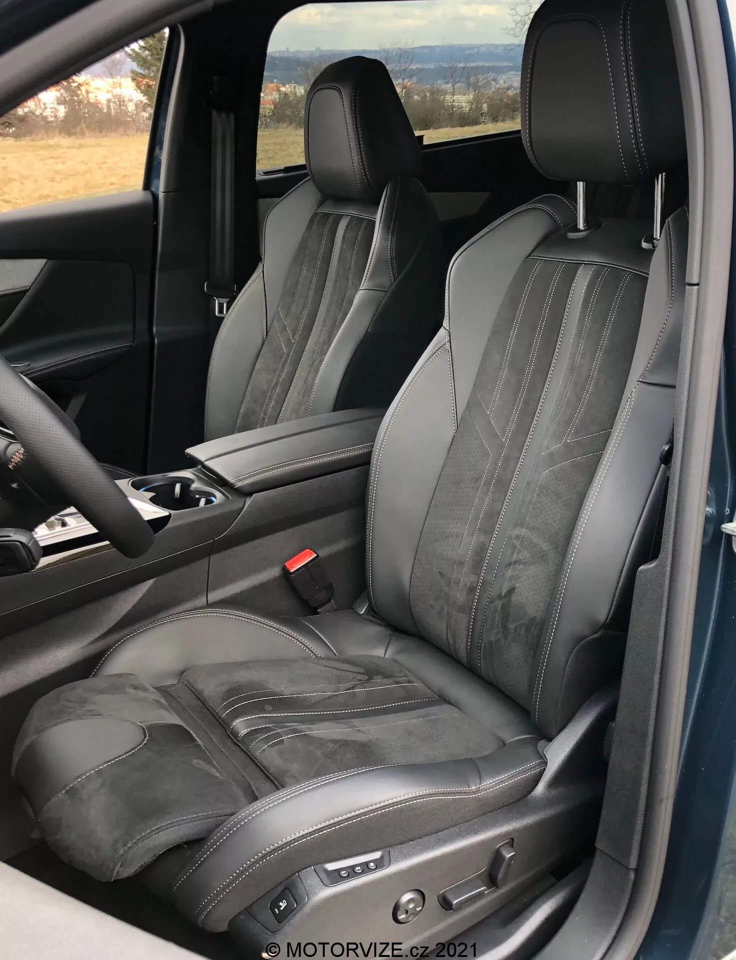 Notranji pogled Peugeot 5008 2021, osredotočen na voznikov sedež, prikazuje nastavljiv črn usnjen sedež s kontrastnim šivom in vodoravnim vtisom, posnet iz perspektive odprtih vrat, poudarjajoč luksuzen in ergonomičen dizajn z elektronskimi nastavitvenimi krmilniki sedeža ter delom sredinske konzole in volana.