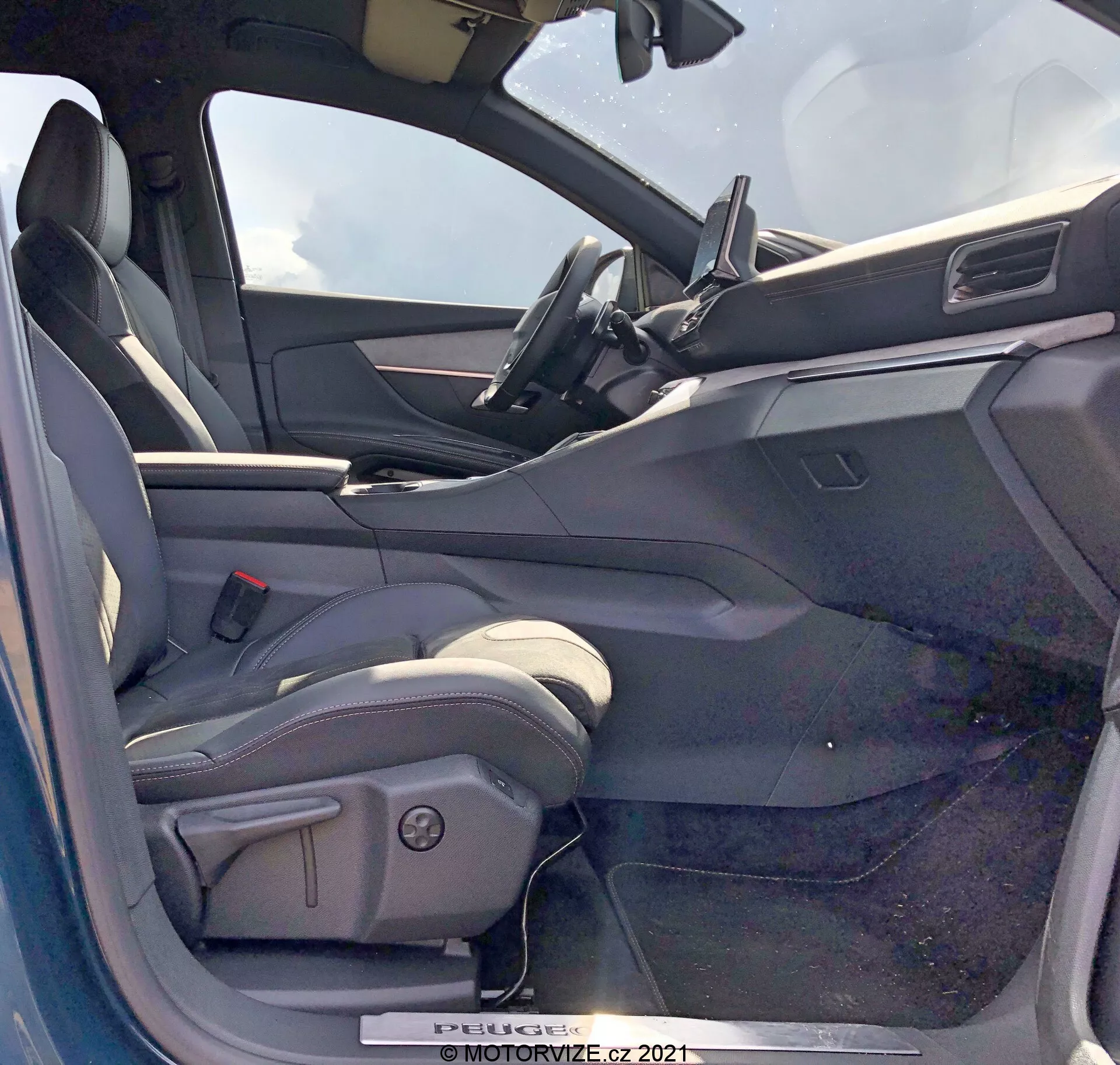 Pogled v notranjost Peugeot 5008 iz leta 2021 s strani voznika z odprtimi vrati. Sedež voznika je v obliki vedra, oblazinjen verjetno v usnje ali usnju podoben material, z bočnimi oporami in električnimi nastavitvenimi kontrolami. Odprta vrata na voznikovi strani prikazujejo notranjo ploščo s kontrolami za električna stekla, verjetno tudi ogledala in mehanizme za zaklepanje vrat. Armatura vsebuje centralno nameščen zaslon na dotik za infotainment z minimalističnim dizajnom. Zgornji del volana je viden, kar nakazuje na volan, oblečen v usnje, z nameščenimi kontrolami. Notranjost ima dvotonsko barvno shemo s temnejšimi zgornjimi površinami in svetlejše spodnje površine.