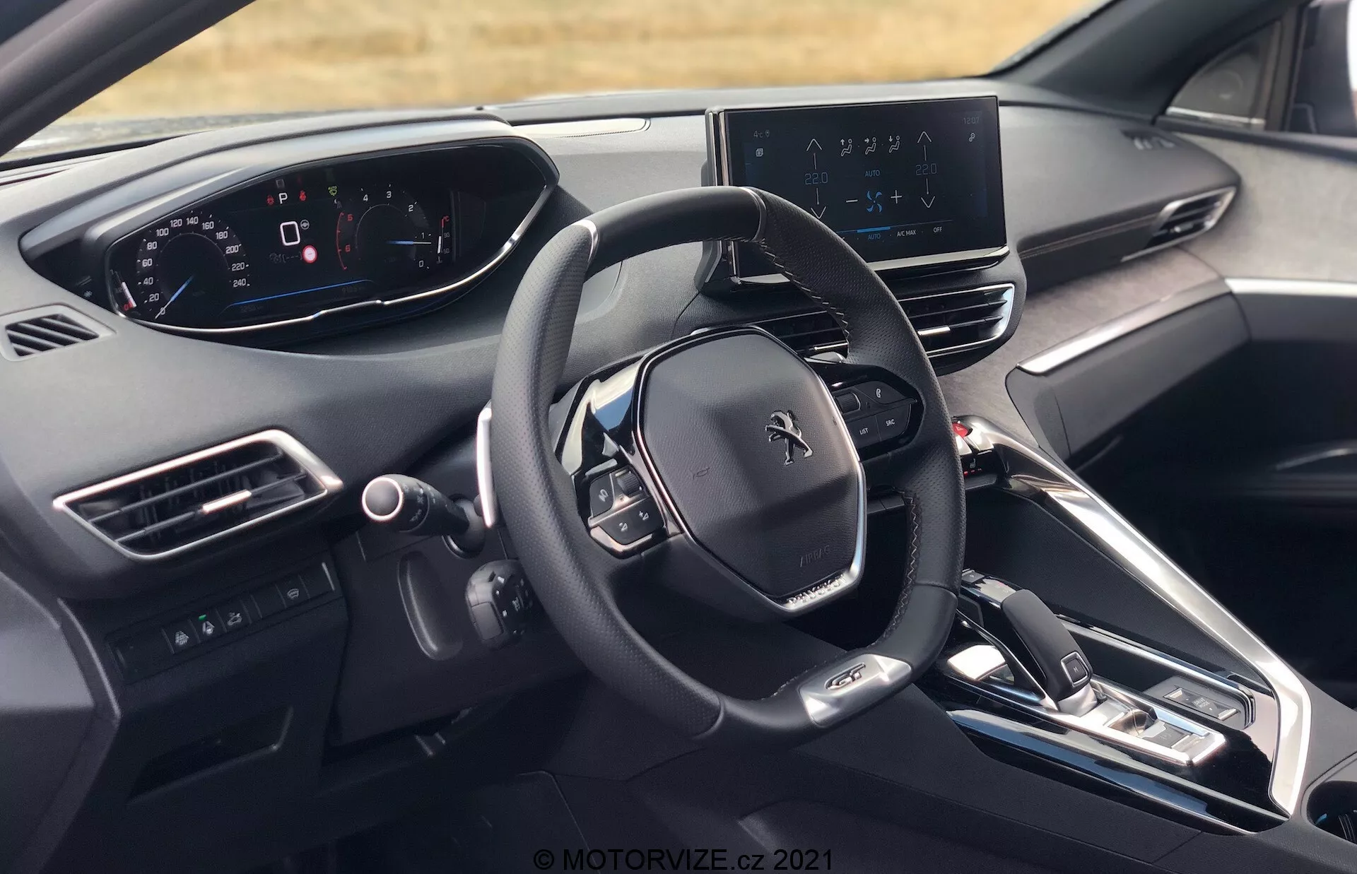 Notranji pogled Peugeot 5008 2021 z voznikove perspektive, ki prikazuje volan z logotipom Peugeot, več kontrolnih gumbov, popolnoma digitalni instrumentni zaslon, osrednji infotainment zaslon z navigacijskim zemljevidom, elegantno armaturno ploščo z minimalnim številom fizičnih gumbov, sredinsko konzolo z gumbi za nadzor klimatskih naprav in nastavitev vozila, elektronsko prestavno ročico, zračnike, gumb za zagon/ustavitev motorja, materiale visokega razreda in delni pogled na vzglavnik voznikovega sedeža.