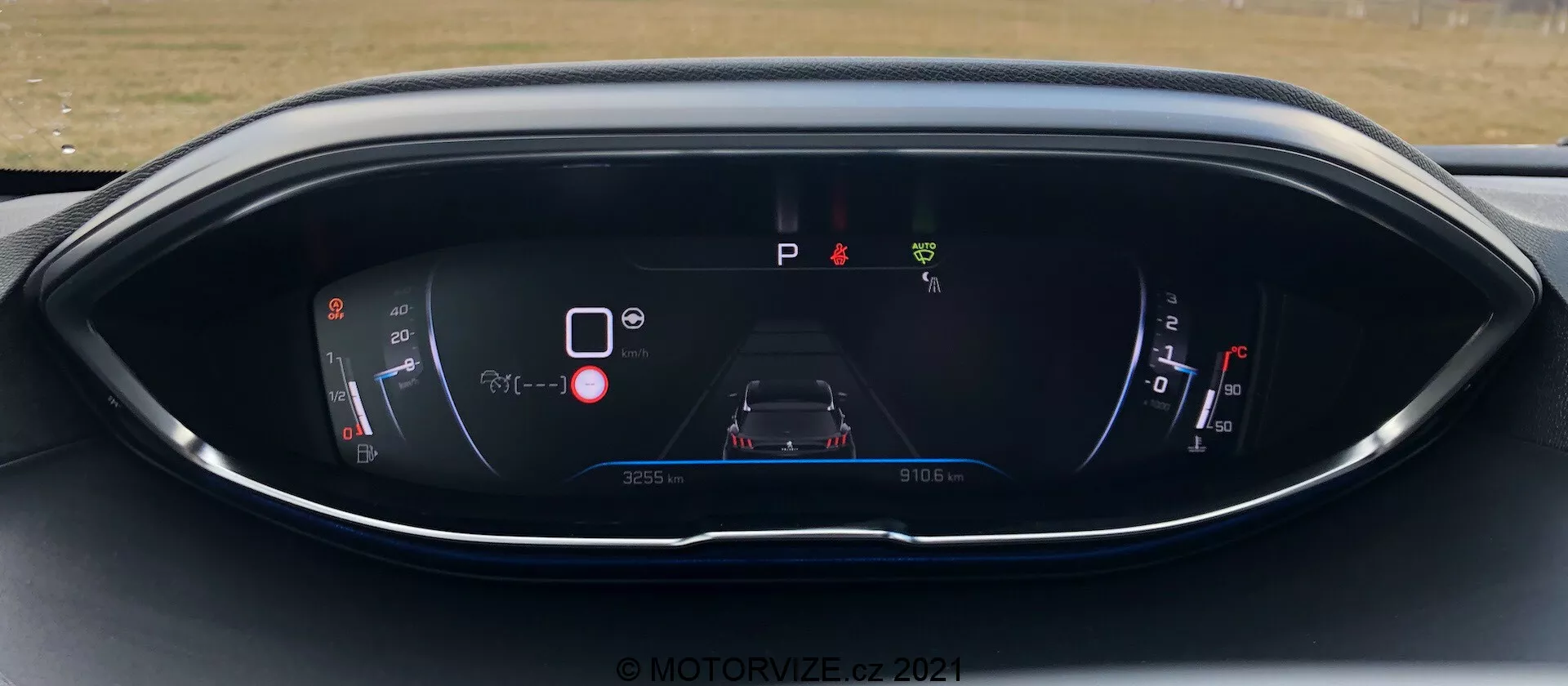 Armaturna plošča Peugeot 5008 2021 s prikazom digitalnega zaslona iz perspektive vozniškega sedeža. Zaslon vključuje kazalnik moči/baterije, osrednji grafični prikaz avtomobila z opozorilnimi ikonami, indikator parkiranja z oznako 'P', kazalnik goriva in informacije o prevoženih kilometrih/potovanju. Oblikovanje je moderno z modro-rdečo barvno shemo na elegantnem, ukrivljenem ohišju. Vodni žig © MOTOR1ZED 2021.