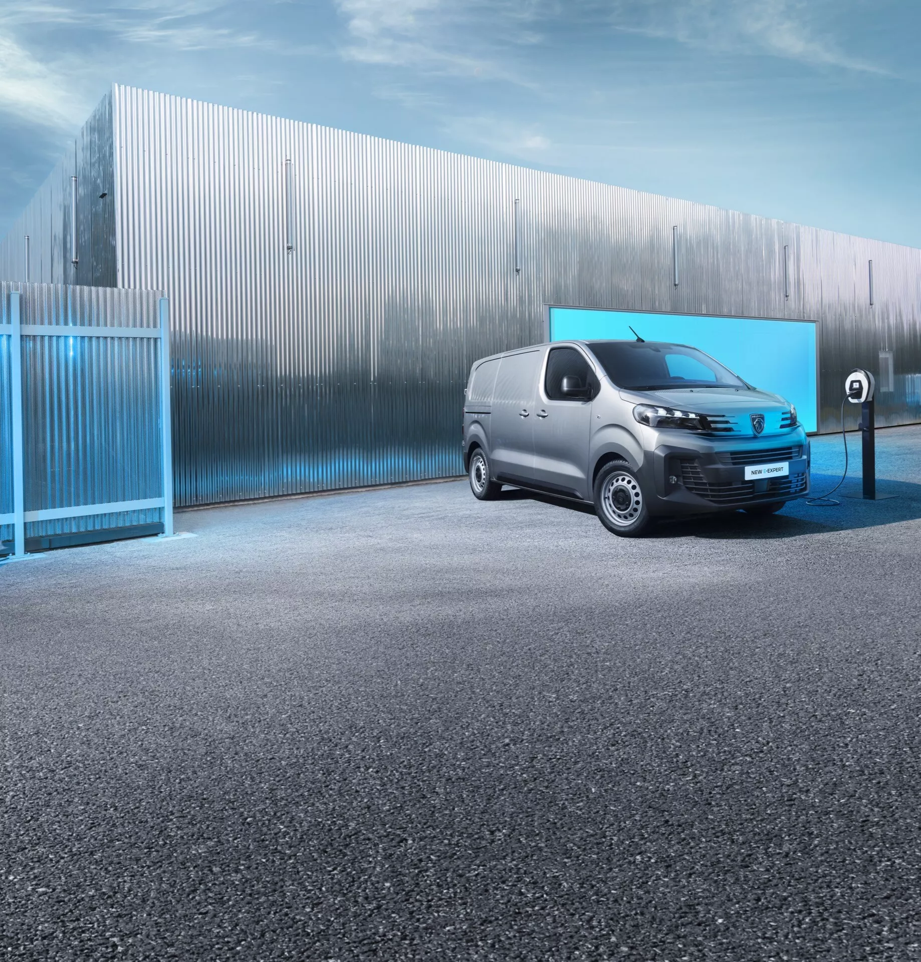Električni dostavnik Peugeot Expert 3. generacije po prenovi 2024 v pogledu s treh četrtin spredaj, poudarjajoč LED žaromete, minimalno rešetko, odprt polnilni vhod, na tlakovani površini z industrijskim ozadjem