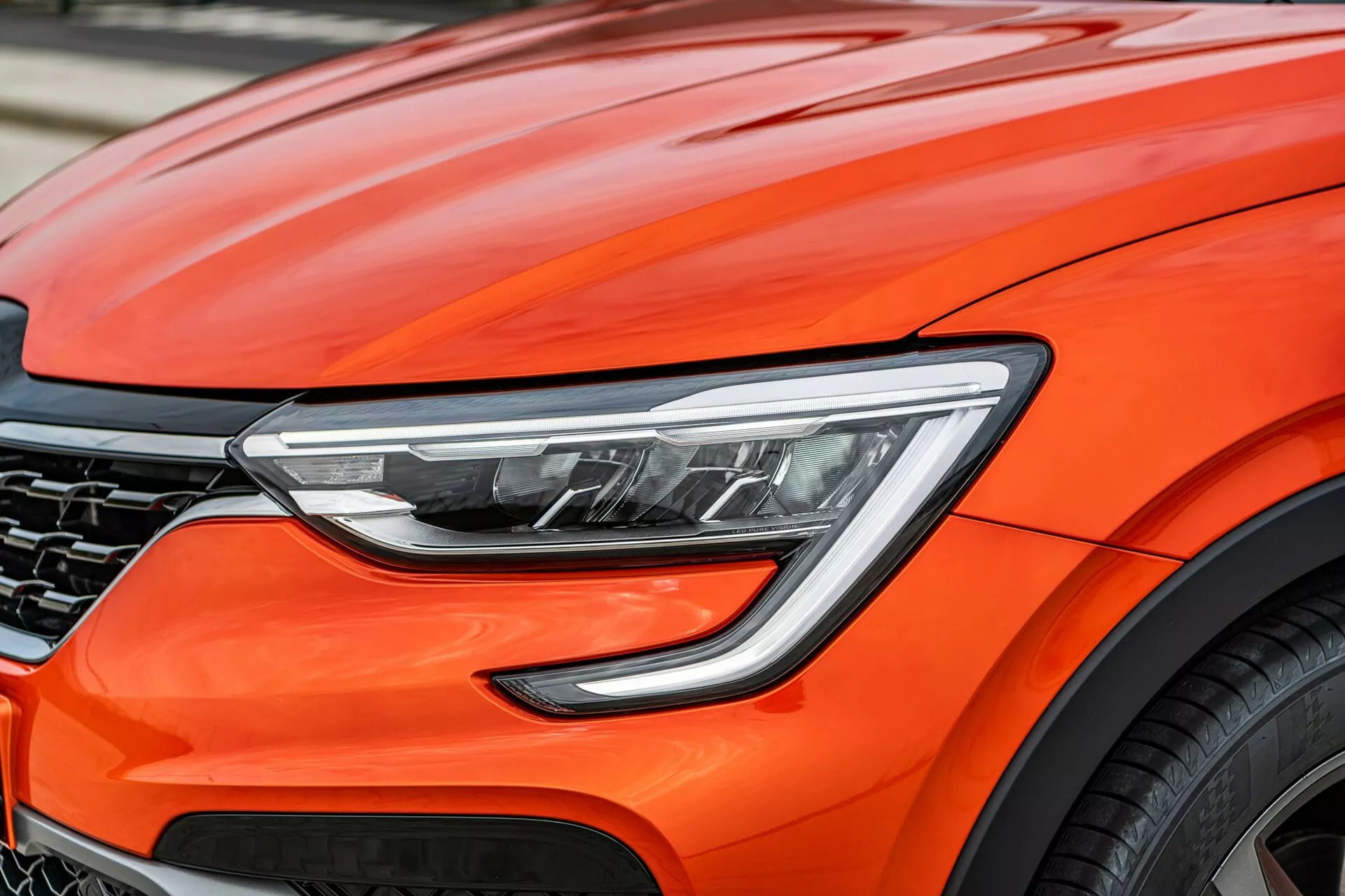 Od blizu zajet pogled na sprednji del svetlo oranžnega Renaulta Arkana 2021, s poudarkom na modernih oblikovalskih detajlih, vključno z naprednimi LED žarometi, aerodinamičnimi linijami pokrova motorja, delno vidno temno mrežasto masko in delom velike aluminijaste kolesne, kar namiguje na vrhunske zmogljivosti in napredno avtomobilsko tehnologijo.
