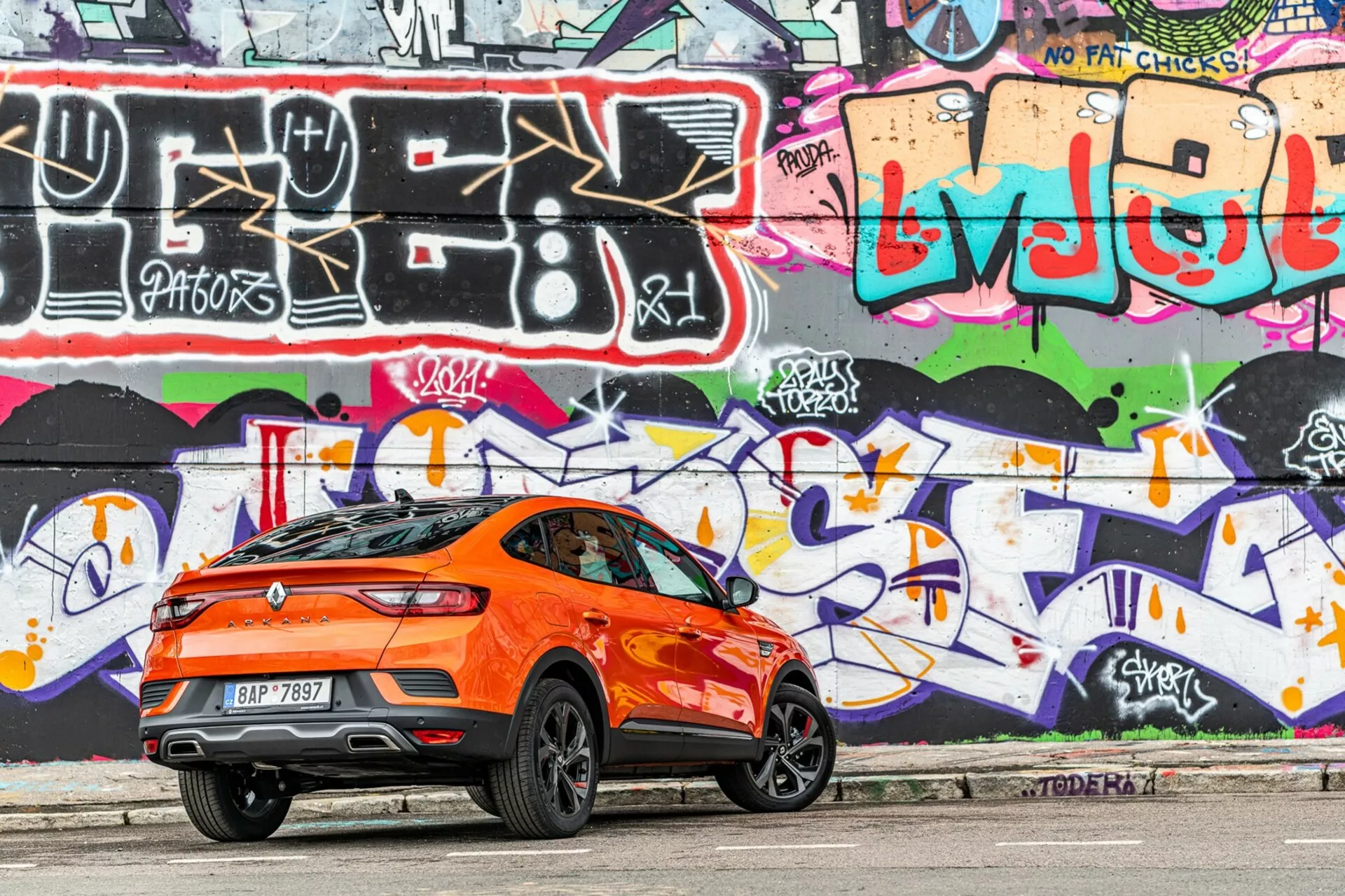 Stranski profil svetlo oranžnega crossoverja Renault Arkana 2021, parkiranega na mestni cesti. Fotografirano s desne strani s poudarkom na zadnji tretjini vozila, slika zajame športno nagnjeno linijo strehe, zadnji spojler, visoko pasno linijo, velika stilska aluminijasta platišča, zatemnjena okna in aerodinamične karakterne linije. Zadnje luči so elegantne in verjetno LED. V ozadju je živahna stena poslikana z grafiti, kar izboljša urbano estetiko. Fotografija je posneta pri dnevni svetlobi in prikazuje mehke sence pod avtomobilom, kar izpostavlja podrobnosti dizajna.