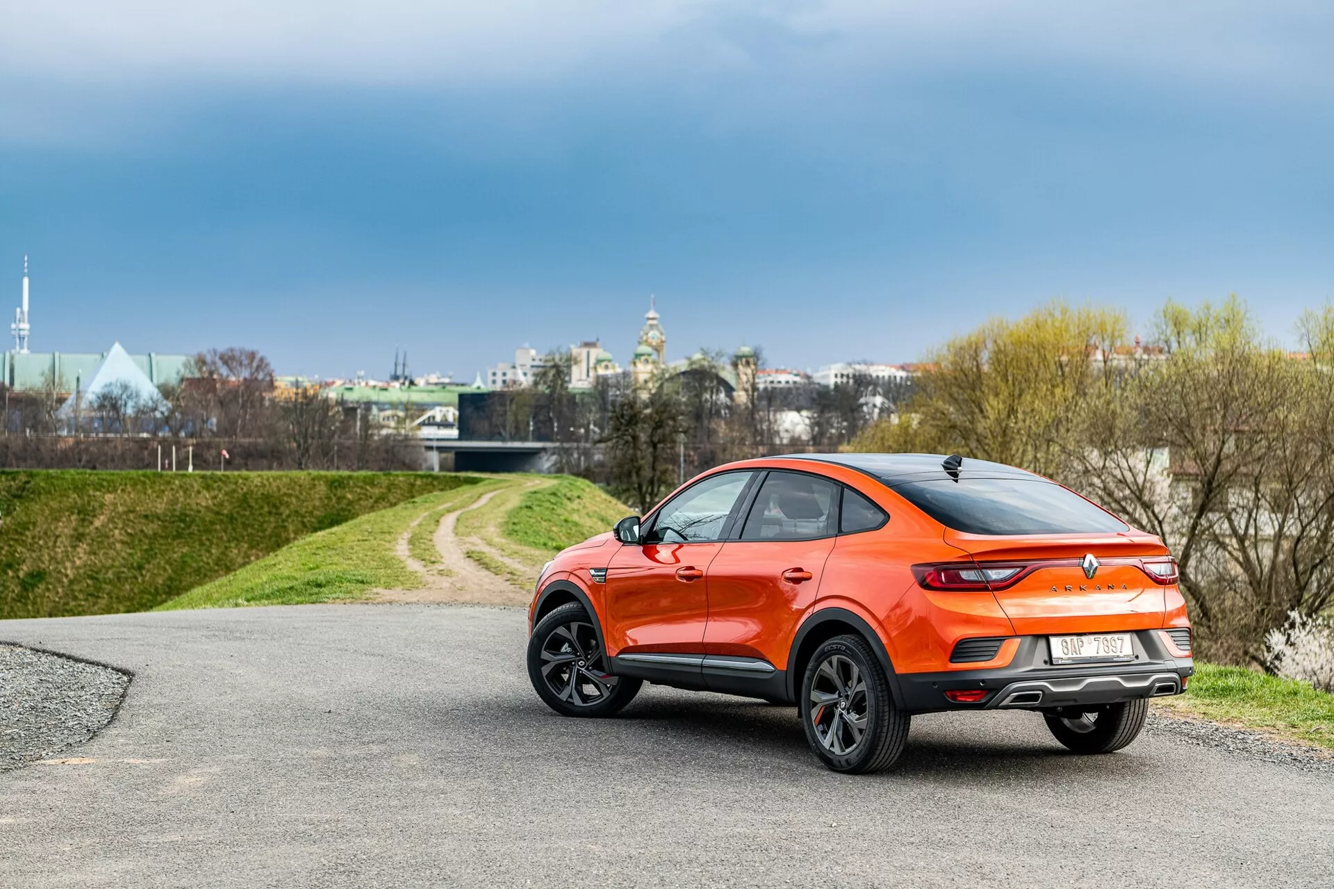 Pogled s treh četrtin od zadaj na hatchback Renault Arkana 2021 s športnim crossover dizajnom v živahni oranžni kovinski barvi. Avto ima nagnjeno streho, črne plastične obloge okoli blatnikov, spojler na vrhu vrat prtljažnika, elegantne LED zadnje luči in velika večkraka lita platišča. Postavljen je na asfaltirani cesti, ki vodi do travnatega območja, pod oblačnim nebom v bližini urbanega, vendar mirnega okolja.