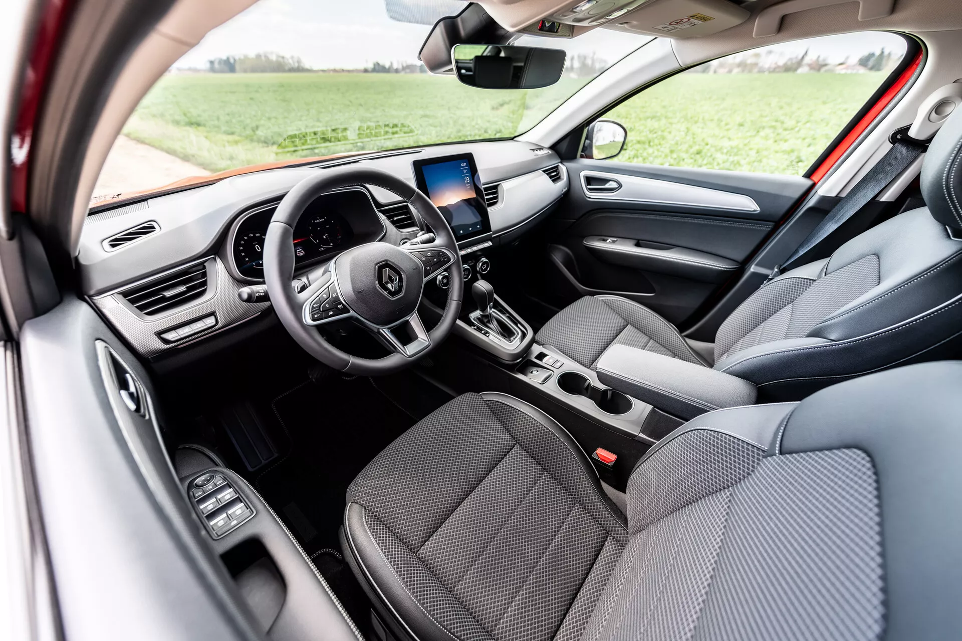 Pogled na notranjost Renault Arkana 2021 s sprednjima sedežema, oblečenima v sivo tkanino, večnamenskim volanom s tremi kraki z vgrajenimi kontrolnimi gumbi, digitalnim prikazovalnikom na armaturni plošči, zaslonom za infotainment z navigacijo GPS nad sredinskimi prezračevalnimi odprtinami, ročnimi upravljalniki za klimatizacijo, avtomatsko prestavno ročico in kontrolniki za električna okna na vratih voznikove strani, osvetljeno z naravno dnevno svetlobo v zunanjem okolju