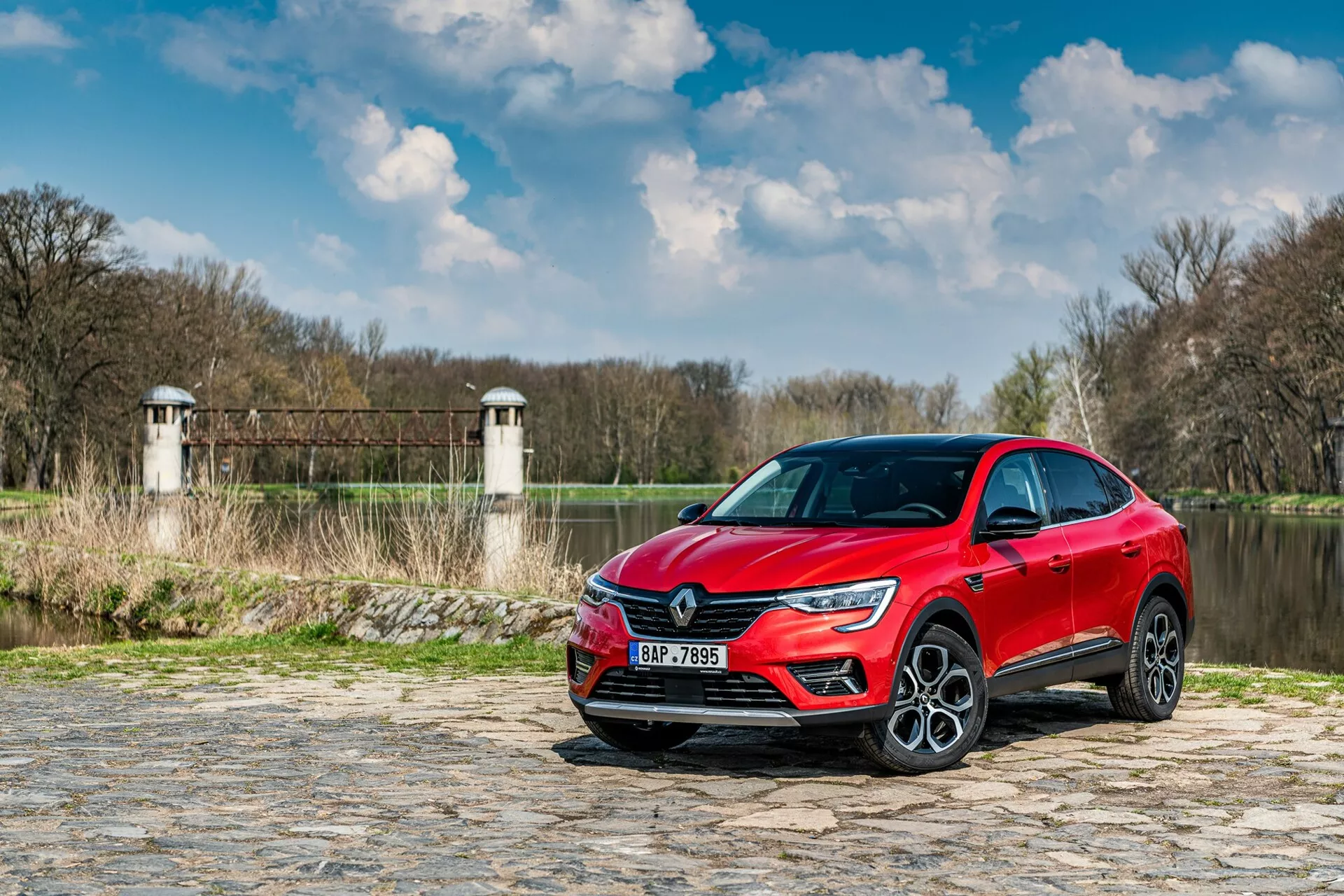 Slika prikazuje Renault Arkana 2021, parkiran na tlakovani poti blizu reke ali jezera, z vegetacijo in zgradbami podobnimi stolpom jezu ali mosta v ozadju. Avto je ujet iz nizkega kota, tričetrtinski pogled spredaj, poudarjajoč sprednjo masko, LED žaromete in del stranskega profila vključno z voznikovimi vrati in sprednjim blatnikom. Sodobni oblikovni elementi, kot so ostra svetlobna podpis LED in velika lita kolesa, kažejo na višjo raven opreme. Reflektivna kovinska barva dodaja estetski pridih, medtem ko možni senzorji za pomoč vozniku in tehnologija, vgrajena v odbijače in stranska ogledala, povečujejo funkcionalnost.