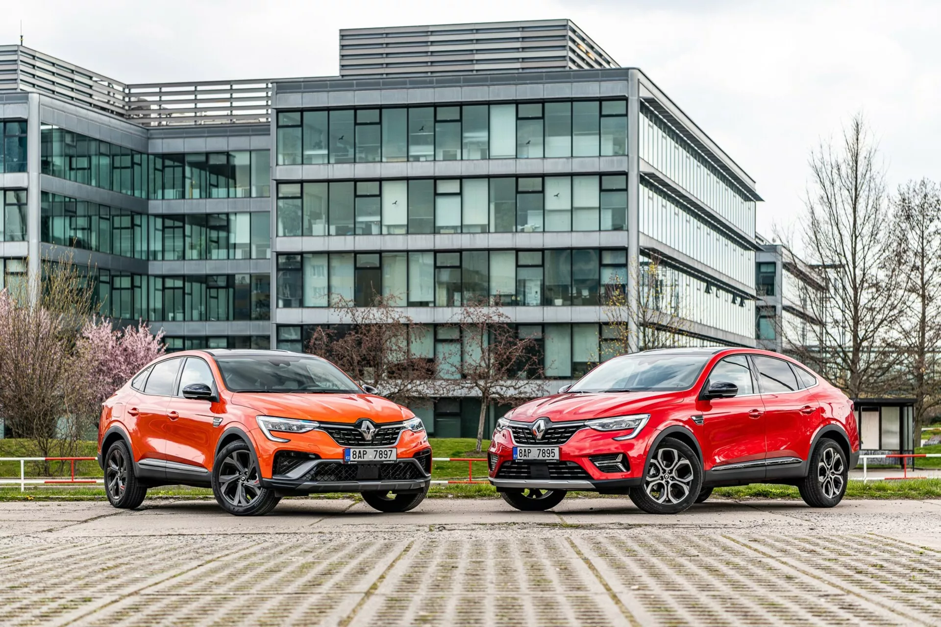 Slika prikazuje dve SUV vozili parkirani drugo ob drugem pred sodobno pisarniško stavbo s stekleno fasado. Vozilo na levi je pobarvano v živahno oranžno barvo z kovinskim sijajem in vsebuje značilno sprednjo mrežo, ostre LED žaromete, izklesano pokrov motorja in velika večkraka aluminijasta platišča z nizkoprofilnimi pnevmatikami. Avto na desni ima kovinsko rdečo barvo, s podobno silhueto, ostrimi žarometi, izrazito mrežo in elegantnimi velikimi aluminijastimi platišči. Obe vozili se ponašata z oglato linijo, aerodinamičnimi ogledali in kontrastno spodnjo obrobo. Fotografija, posneta iz nizkega kota blizu nivoja tal, poudarja urbano okolje in izpostavlja obliko, prisotnost in napredne tehnološke funkcije vozil.