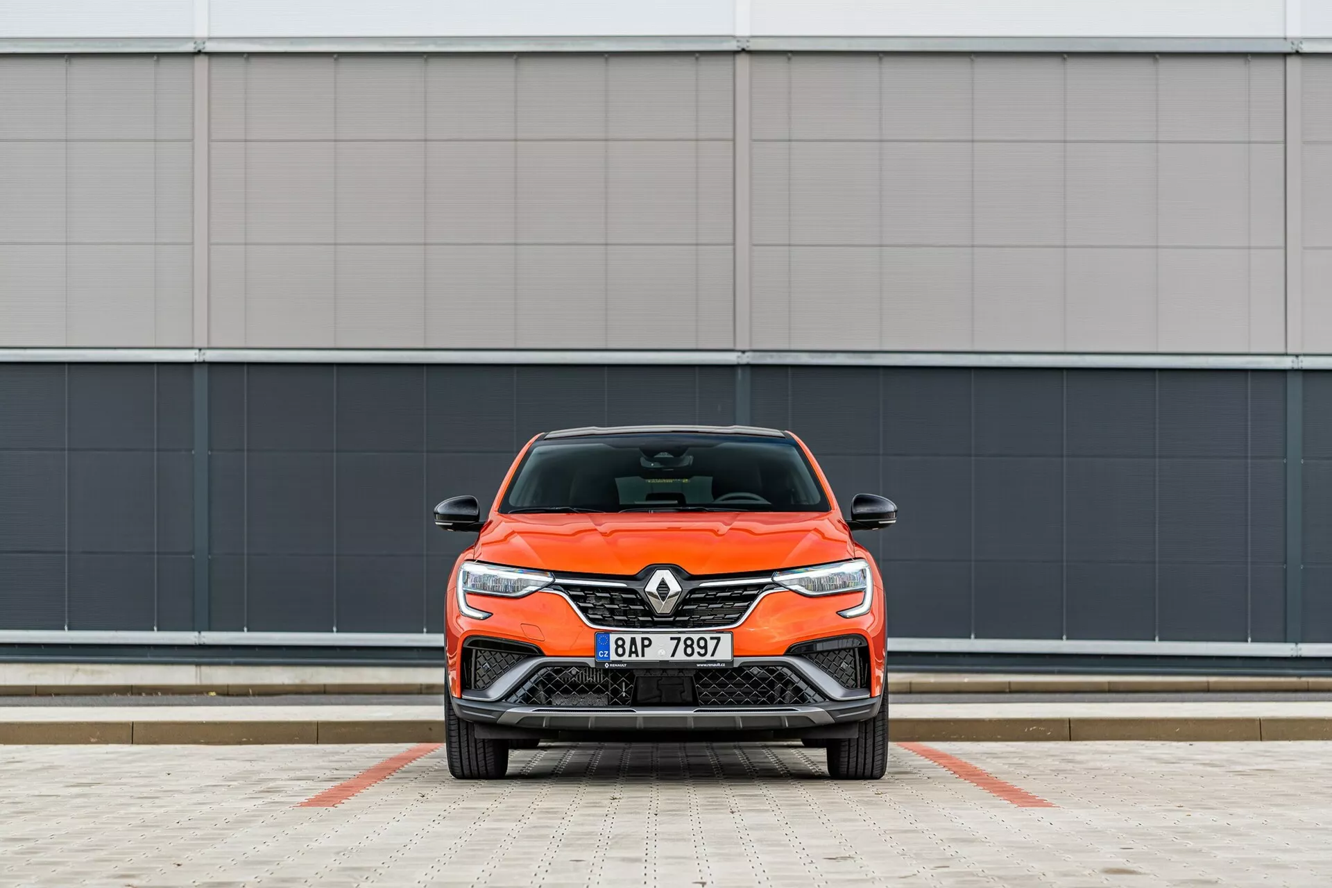 Frontalni pogled na svetlo oranžen Renault avtomobil, ki poudarja njegov sodoben dizajn z elegantnimi žarometi in prednjo masko, parkiran pravokotno na kamero ob minimalistični sivi steni v označenem parkirnem prostoru.