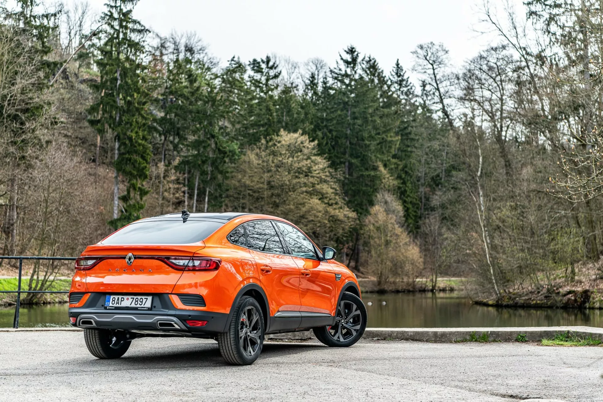 Renault Arkana 2021 prikazan pod kotom treh četrtin od zadaj, podroben pogled na stranski in zadnji profil, prikazuje sodoben crossover/SUV z dvignjeno oddaljenostjo od tal in športno oblikovano streho v slogu kupeja. Živahna oranžna karoserija izstopa v ozadju dreves in jezera, poudarjajoč večkraka aluminijasta platišča, zadnje LED luči, zadnji spojler, anteno v obliki morskega psa, sredinsko nameščen emblem, dvojne izpušne cevi, sijoč zaključek laka in zadnja zatemnjena stekla. Razpršena naravna osvetlitev izboljša vidljivost dobro vzdrževane zunanjosti, kar nakazuje privlačnost za potrošnike z aktivnim življenjskim slogom, parkirano na tlakovani površini.