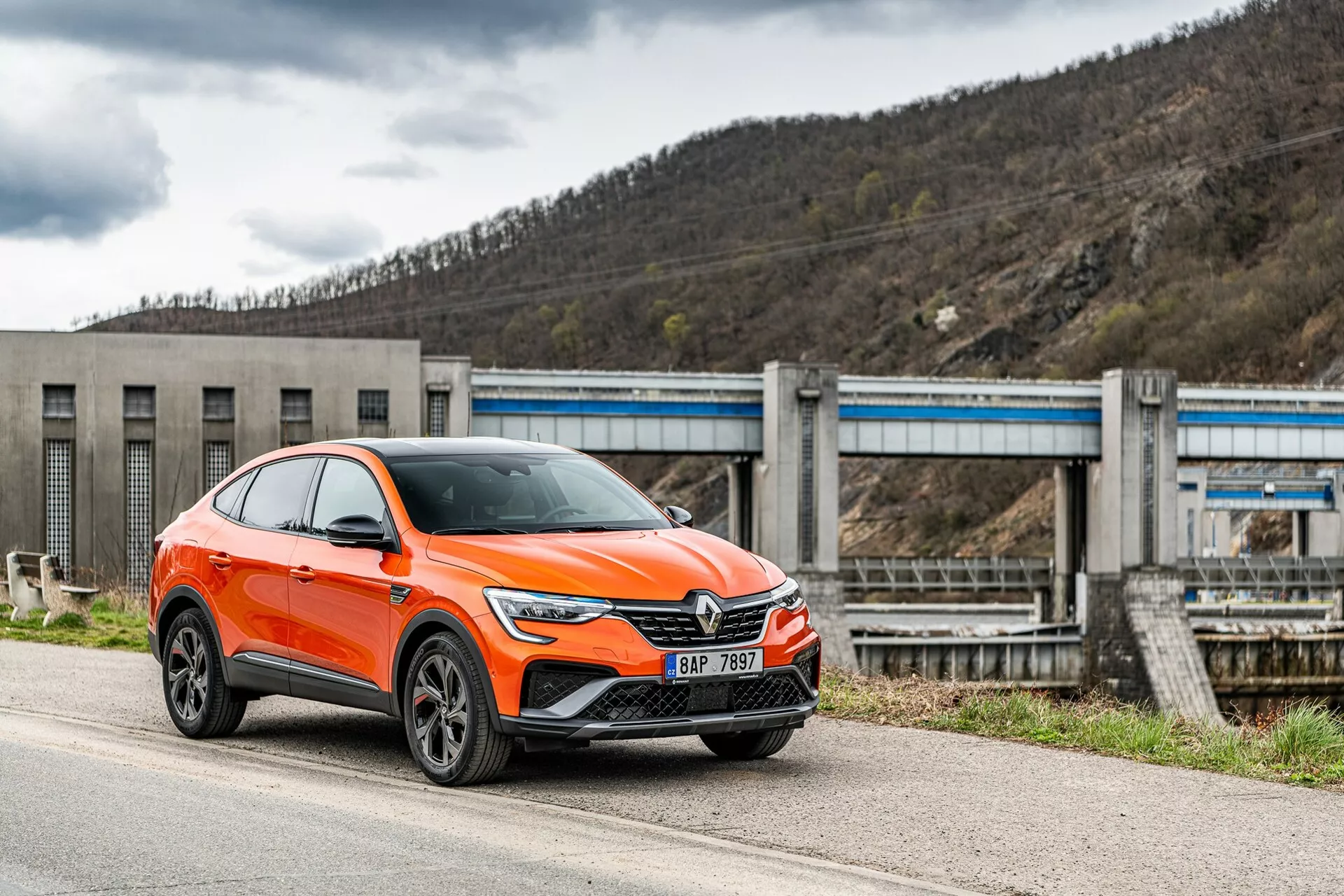 Moderen svetlo oranžen crossover SUV Renault Arkana 2021 je parkiran ob cesti, fotografiran iz spredaj-sstranskega kota, osredotočenega na voznikovo stran. Slika prikazuje sprednjo masko avtomobila z velikim emblemom, elegantne kotne LED žaromete, dnevne luči, zračne dovode in verjetno senzorje asistenčnih sistemov za voznika na spodnjem odbijaču, izrazite blatnike s platišči iz lahke litine in zrcala z integriranimi smerniki. Vozilo, ki je razvrščeno med SUV-je, ima povišana stojišča z večjo oddaljenostjo od tal in rahlo nagnjeno streho v stilu kupeja. V ozadju je vidna infrastruktura, ki spominja na jez ali sistem zapornic, ki postavlja vozilo v industrijsko ali podeželsko okolje. Posnetek je narejen iz rahlo nizkega kota, kar poudarja višino in stojišče avtomobila.
