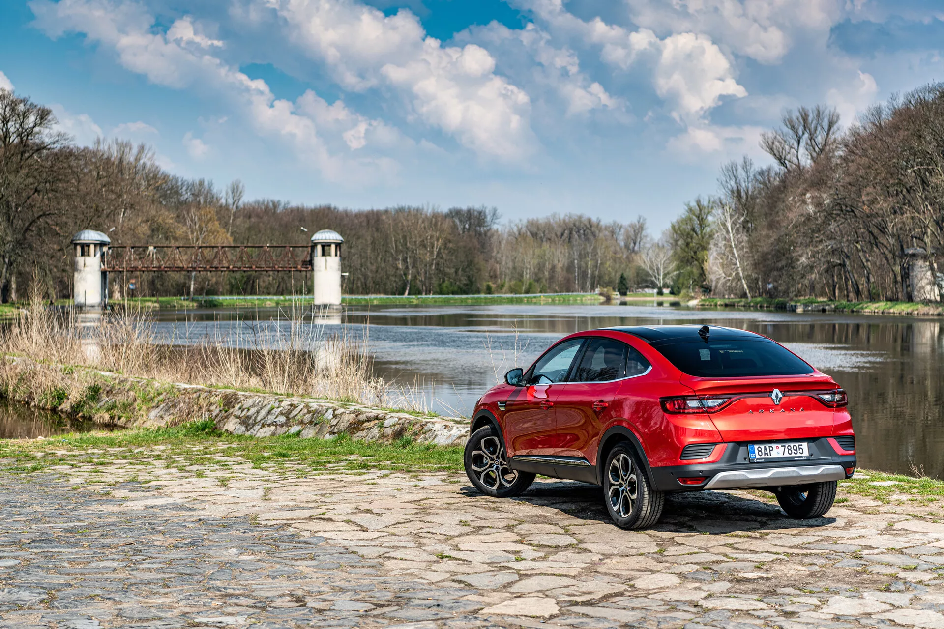 Renault Arkana 2021, parkirana na tlakovcu blizu vode z naravnim ozadjem, ki prikazuje zadnji tričetrtinski pogled, športni rdeč dizajn in LED zadnje luči