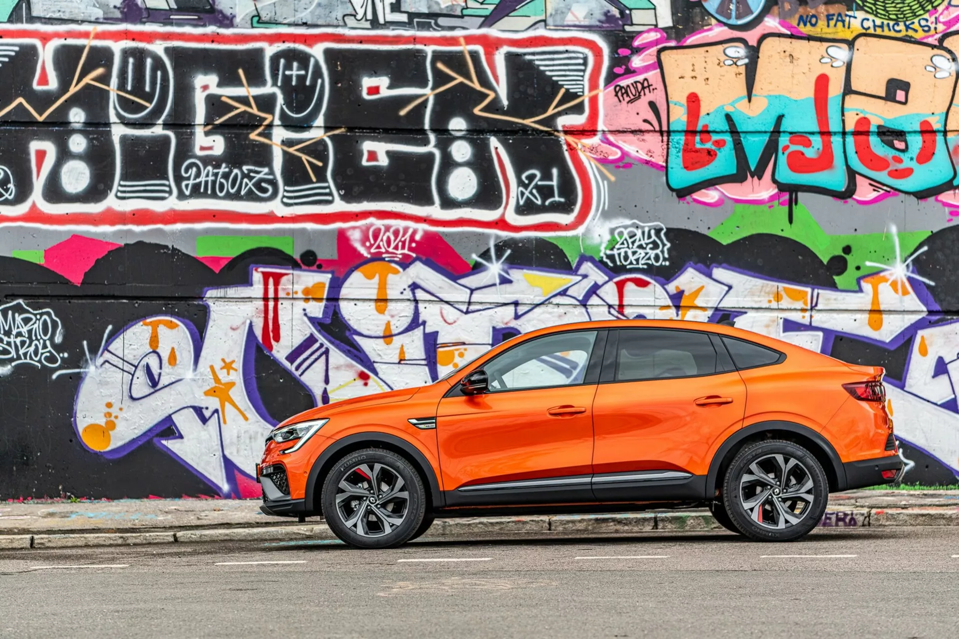 Slika prikazuje živahno oranžni crossover SUV Renault Arkana 2021, parkiran vzporedno z ulico, z živahno barvito grafitno steno v ozadju. Fotografija, posneta iz bočnega kota, poudarja športen in sodoben dizajn vozila z vidnimi voznikovimi prednjimi in zadnjimi vrati, stranskimi okni, stranskimi ogledali in poševno strešno linijo. Stilizirana aluminijasta kolesa z nizkoprofilnimi pnevmatikami nakazujejo osredotočenost na cestno zmogljivost. Ključi za brezkljični vstop na sprednjih vratih nakazujejo brezkljični vstopni in zagonski sistem. Slika je posneta na dnevni svetlobi, poudarja kontrast oranžne karoserije proti grafitu, prikazuje dobro vzdrževano, ostro karoserijo vozila v mladostnem, urbanem okolju.