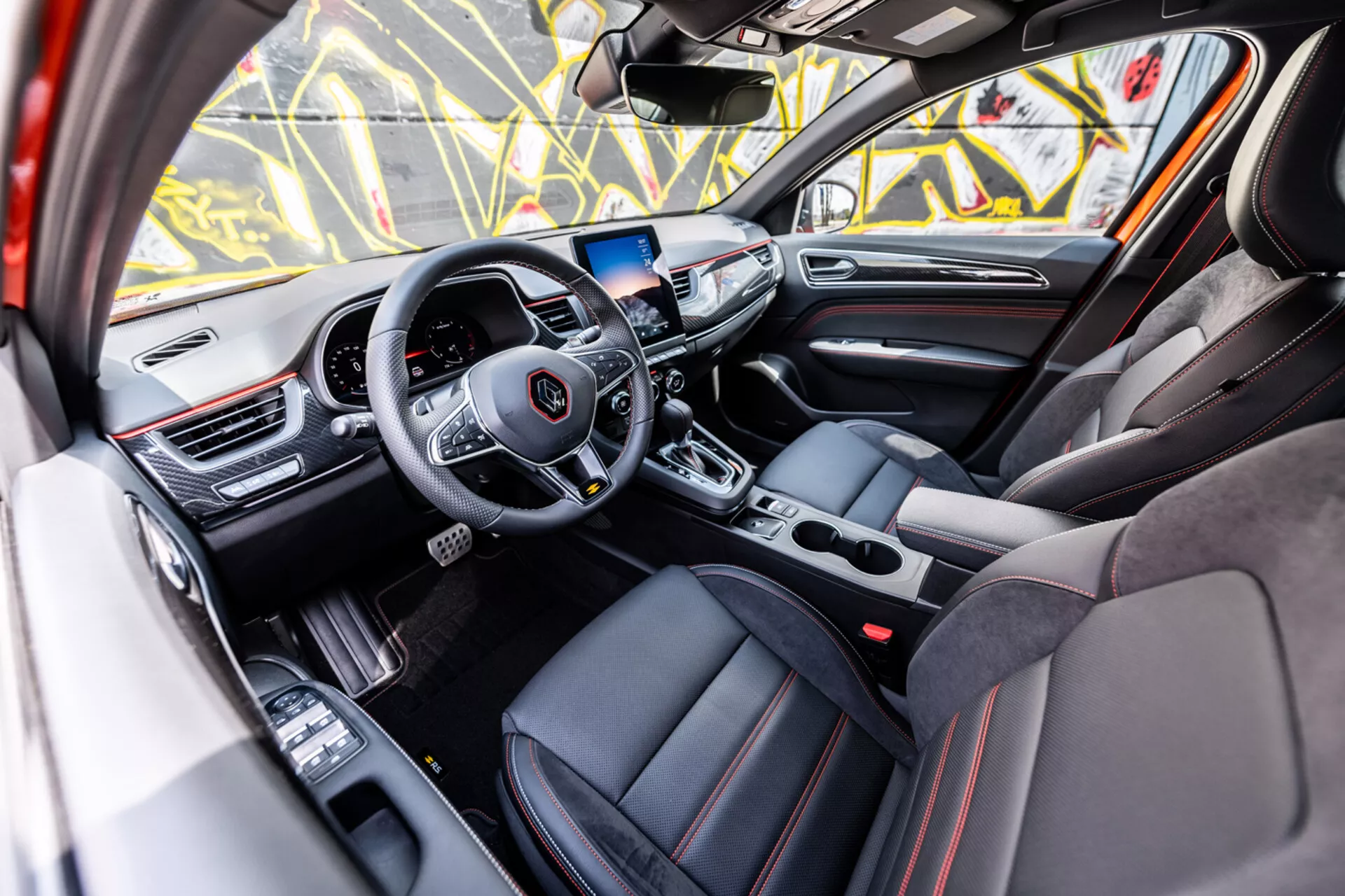 Notranjost Renault Arkana 2021, ki prikazuje razkošno sprednjo kabino iz perspektive vrat sopotnika z modernim volanom s tremi kraki, zaslon na dotik za infotainment in športno oblazinjenje z rdečimi šivi