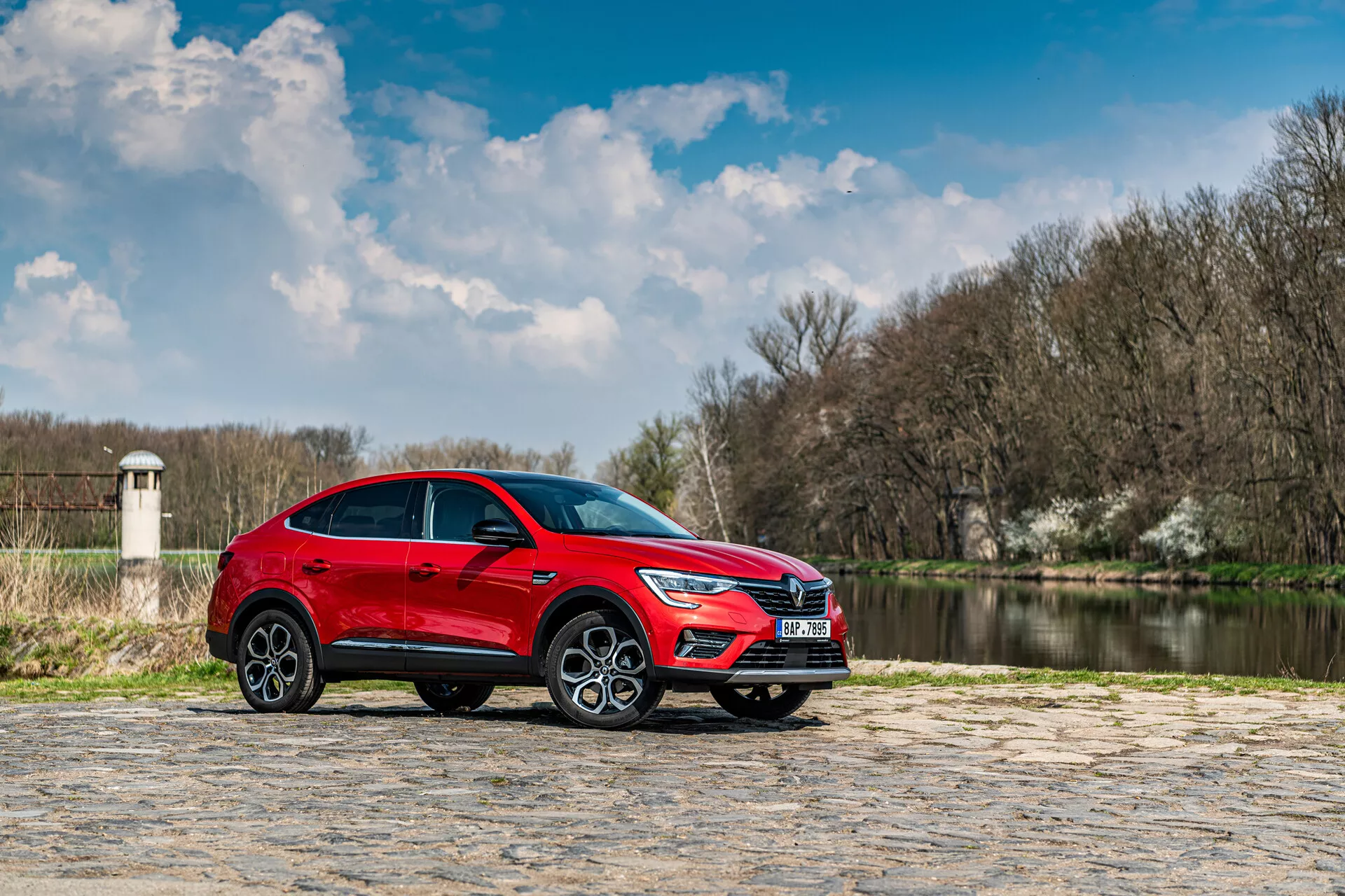 Rdeč SUV Renault Arkana 2021 parkiran na kamniti površini z vodo in drevesi v ozadju pod delno oblačnim nebom, stranski profil poudarja elegantno oblikovanje in dinamične lastnosti