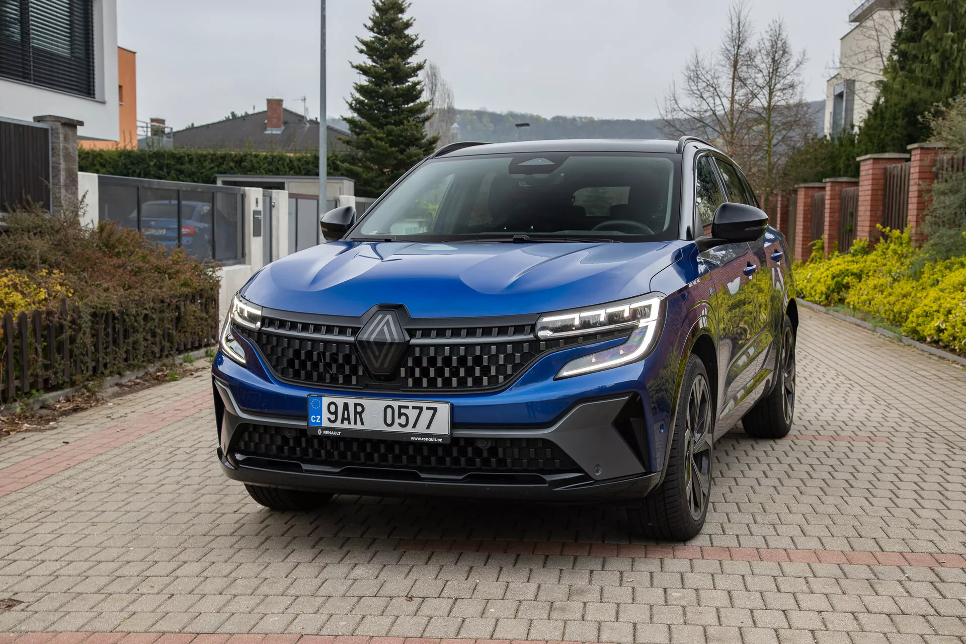 Renault Austral 2022 v modri barvi, prikazuje svoj sprednji tričetrtinski kot na opečnatem dovozu z bivalnim okoljem v ozadju, lastnosti vključujejo LED žaromete, značilno masko, senzorje spredaj, lita kolesa in stranska ogledala z integriranimi smerokazi pod naravno, oblačno dnevno svetlobo.
