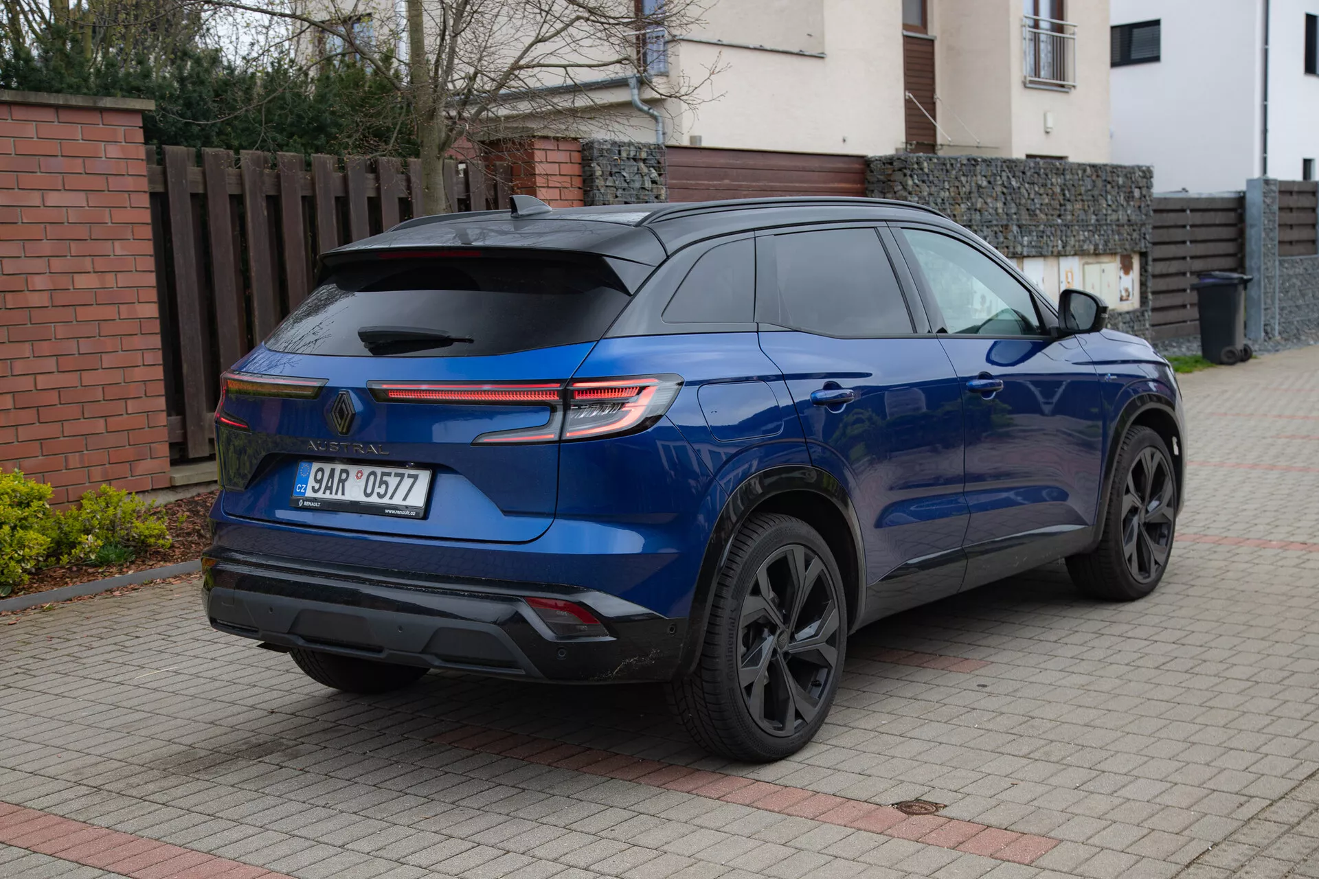 Moderen SUV Renault Austral 2022 v sijoči metalik modri barvi, parkiran na tlakovani dovozni poti ob stanovanjski stavbi, ki prikazuje svoj športni zadnji spojler, LED zadnje luči, temne zlitinske kolesa, zatemnjena zadnja okna in parkirne senzorje iz zadnjega kota treh četrtin