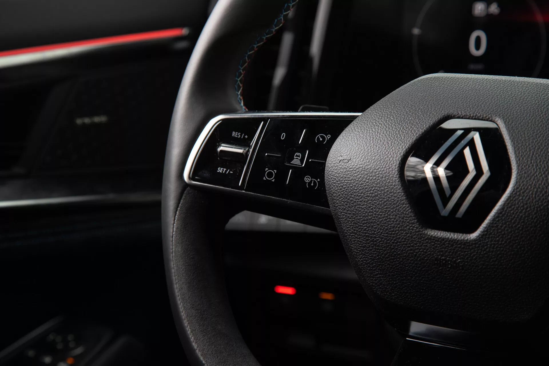 Pogled na notranjost Renault Austral 2022, ki poudarja kontrole na volanu in emblem, vključno z osvetljenimi funkcijami gumbov za glasnost, klice, glasovne ukaze in navigacijo infotainmenta, iz perspektive voznikovega sedeža.