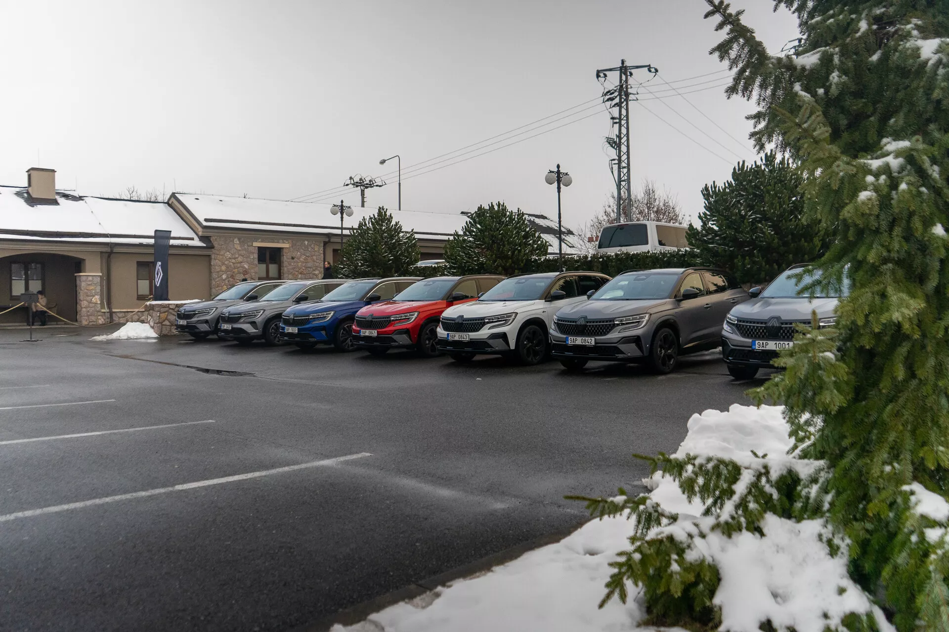 Parkirana vrsta mešanih limuzin in SUV-jev pod lahkim sneženjem, kar nakazuje na hladno vreme, z vidnimi stranskimi profili vključno z vrati, stranskimi ogledali in kolesi, posneto iz dvignjenega kota pločnika