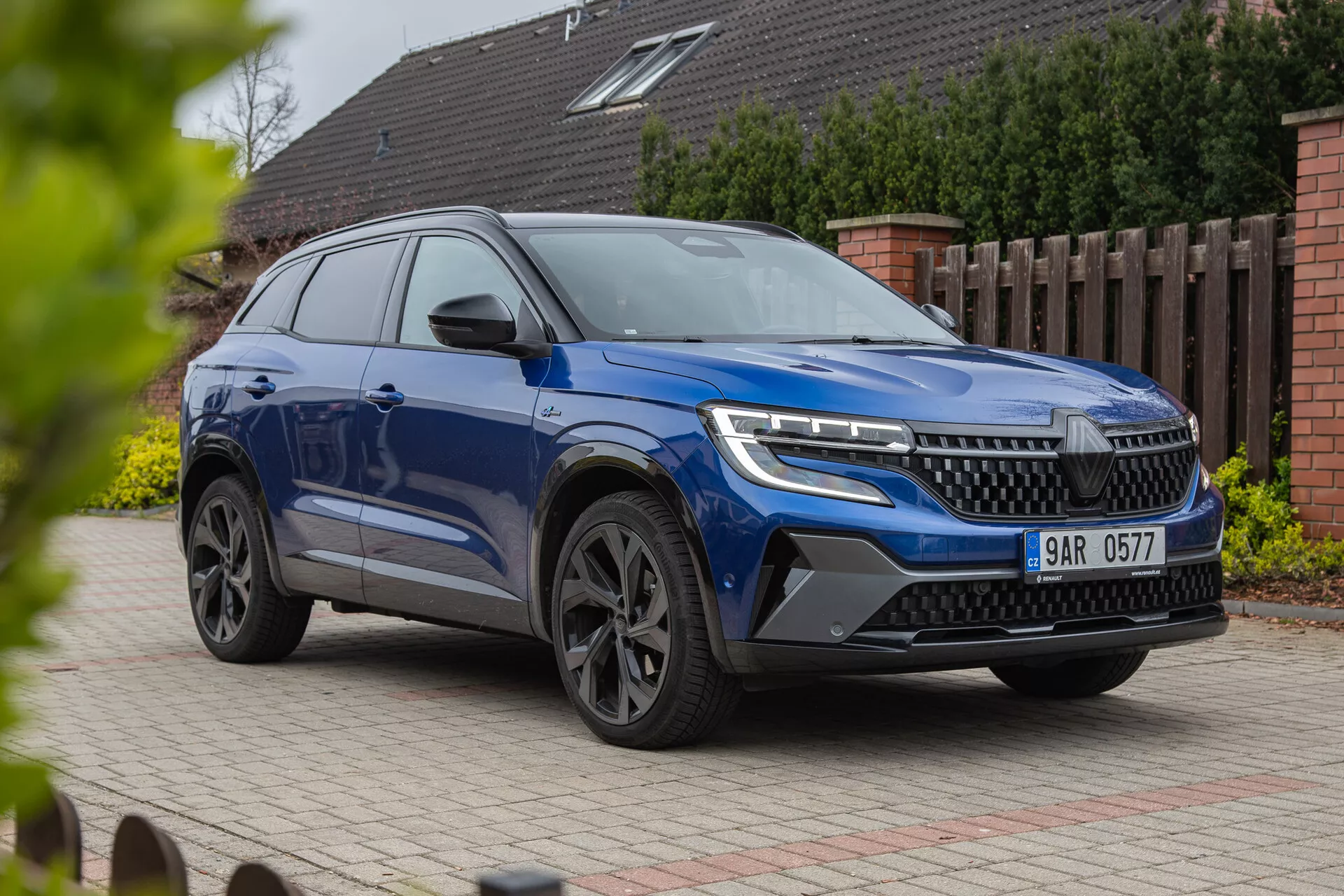 Moderen SUV Renault Austral 2022 prikazan iz tročetrtinskega sprednjega kota, ki poudarja LED žaromete, izstopajočo rešetko, stilna lita kolesa in dinamično karoserijo z metalik končno obdelavo, fotografiran iz nekoliko nižjega kota, da poudari njegovo postavo in oblikovalske značilnosti na stanovanjskem ozadju.