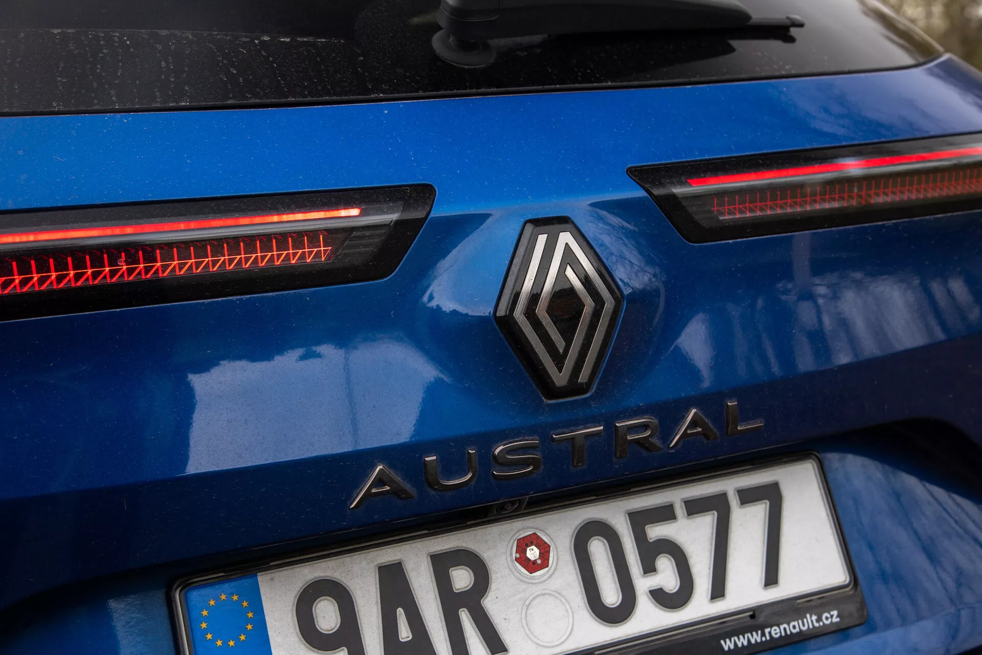 Podroben posnetek zadnjega dela modrega Renault Austral 2022, s centralno postavljenim logotipom v obliki diamanta in imenom modela 'Austral' v velikih črkah, registrsko tablico z zastavo Evropske unije, izstopajočimi vodoravnimi LED zadnjimi lučmi in možnim dodatnim sistemom zadnje razsvetljave, zunanji izgled avtomobila kaže na nedavno uporabo.