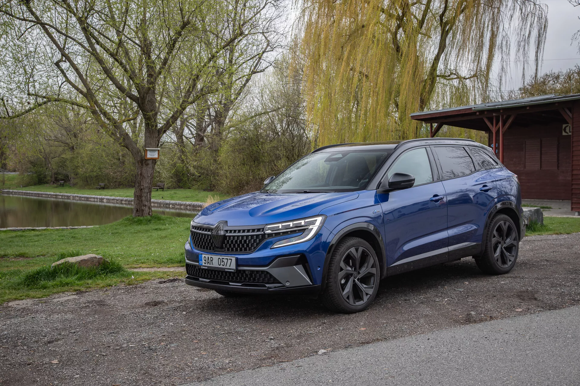 SUV križanec Renault Austral 2022 v metalik modri barvi, parkiran zunaj, prikazuje profil spredaj in s strani z LED žarometi in strešnimi nosilci, obdan z naravnim parkovnim okoljem