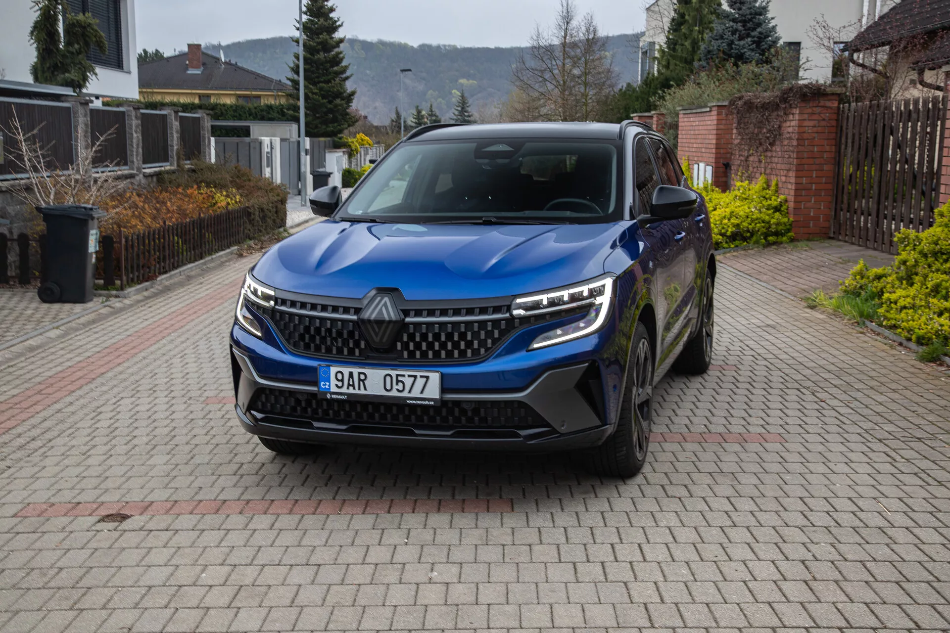 Pogled spredaj pod kotom na SUV Renault Austral 2022 parkiran na dovozni poti v stanovanjski soseski, prikazuje rešetko, LED žaromete, dnevne luči, odbijač z zračnimi vstopi, parkirne senzorje, emblema, lita kolesa, stranska ogledala z vgrajenimi smerokazi in registrsko tablico.