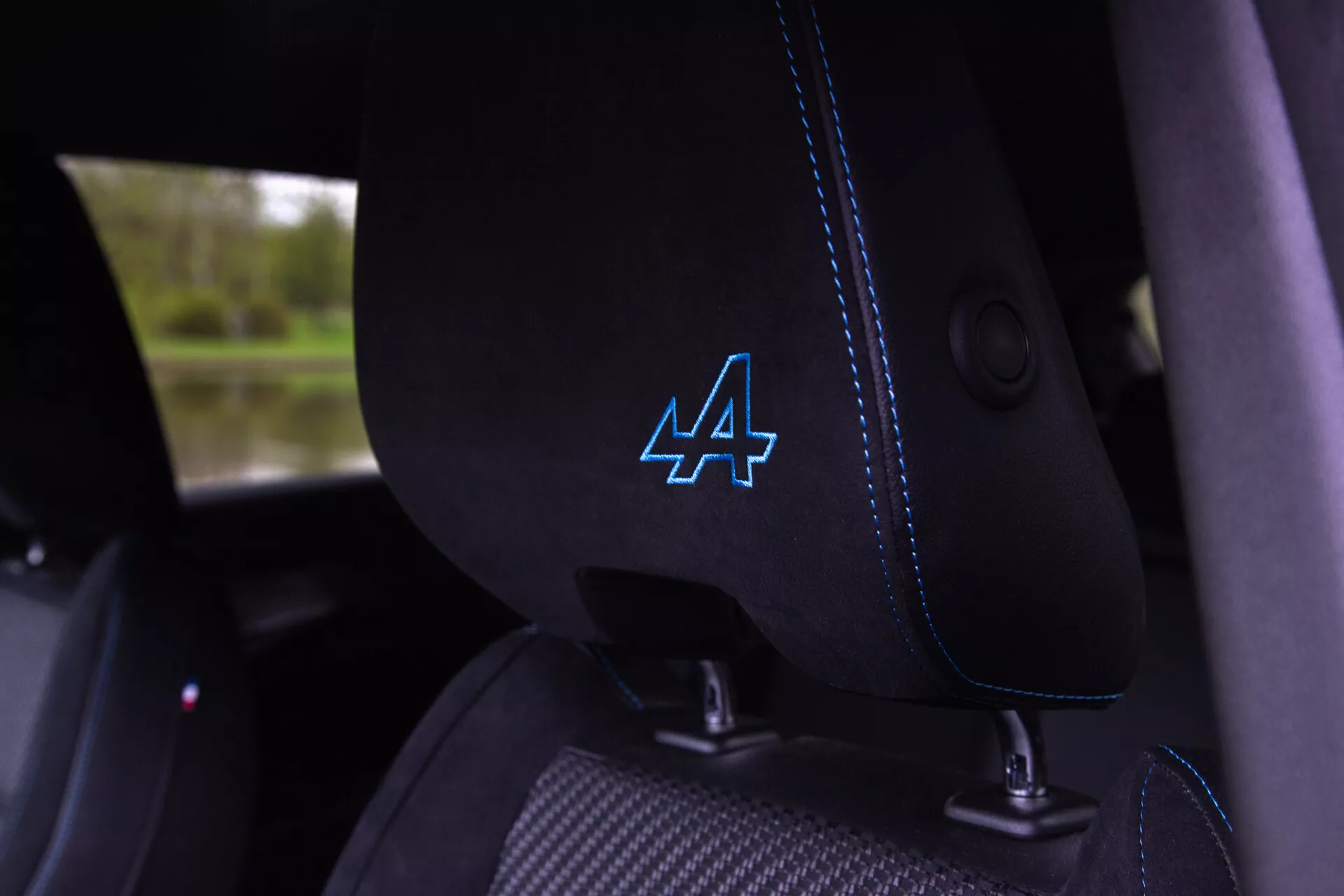 Pogled od blizu notranjosti Renault Austral 2022 prikazuje črni tkaninski voznikov sedež z značilnim modrim šivom in stiliziranim logotipom 'A' na naslonjalu za glavo, viden integriran vodič varnostnega pasu, kar nakazuje sodobne in morda športne značilnosti vozila.