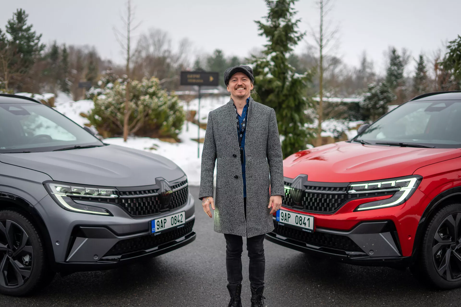 Nasmejana oseba obdana z dvema SUV Renault Austral 2022 v zimski nastavitvi, ki poudarja sodobno zasnovo vozil in napredne tehnološke lastnosti s poudarkom na sprednji rešetki, LED žarometih in elegantnem bočnem profilu.