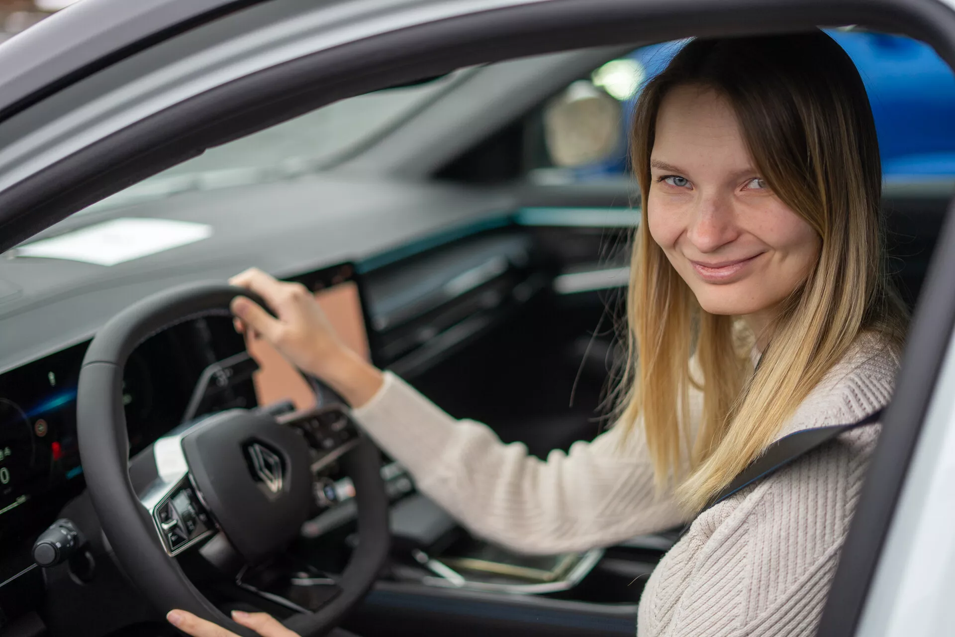 Pogled na notranjost Renault Austral 2022, osredotočen na stran voznika z žensko na voznikovem sedežu, z vgrajenimi kontrolami na volanu, digitalnim instrumentnim zaslonom in centralno konzolo z zaslonskim vmesnikom na dotik.