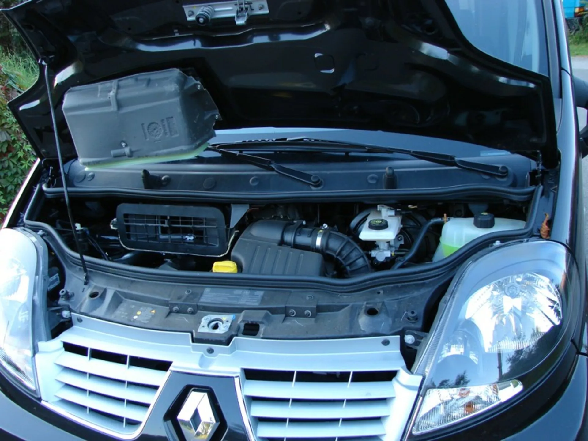 Sprednji tričetrtinski pogled na Renault Trafic Combi 2006 z odprtim pokrovom, ki razkriva motor in komponente, vključno z blokom motorja s pokrovom, zračnim vhodom in rezervoarji za tekočine, obkrožen z tradicionalnimi halogenskimi žarometi