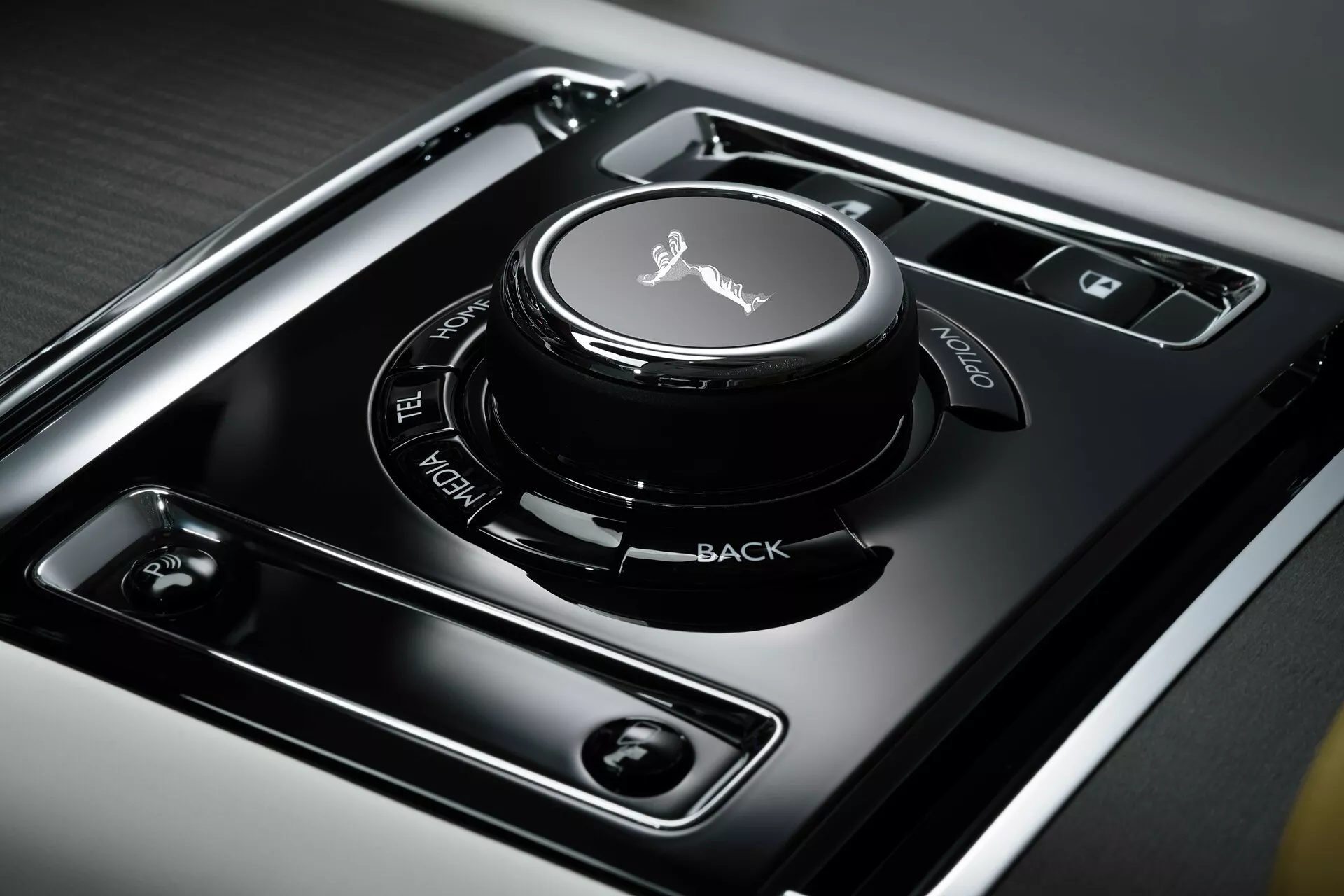 Pogled od blizu na sredinsko konzolo Rolls-Royce Spectre 2023 prve generacije, ki prikazuje gumb za nadzor infotainmenta z občutljivo površino na dotik, obdan z jasno označenimi funkcionalnimi gumbi na visoko sijoči črni plošči, vstavljen v brušen kovin za razkošno notranjo opremo