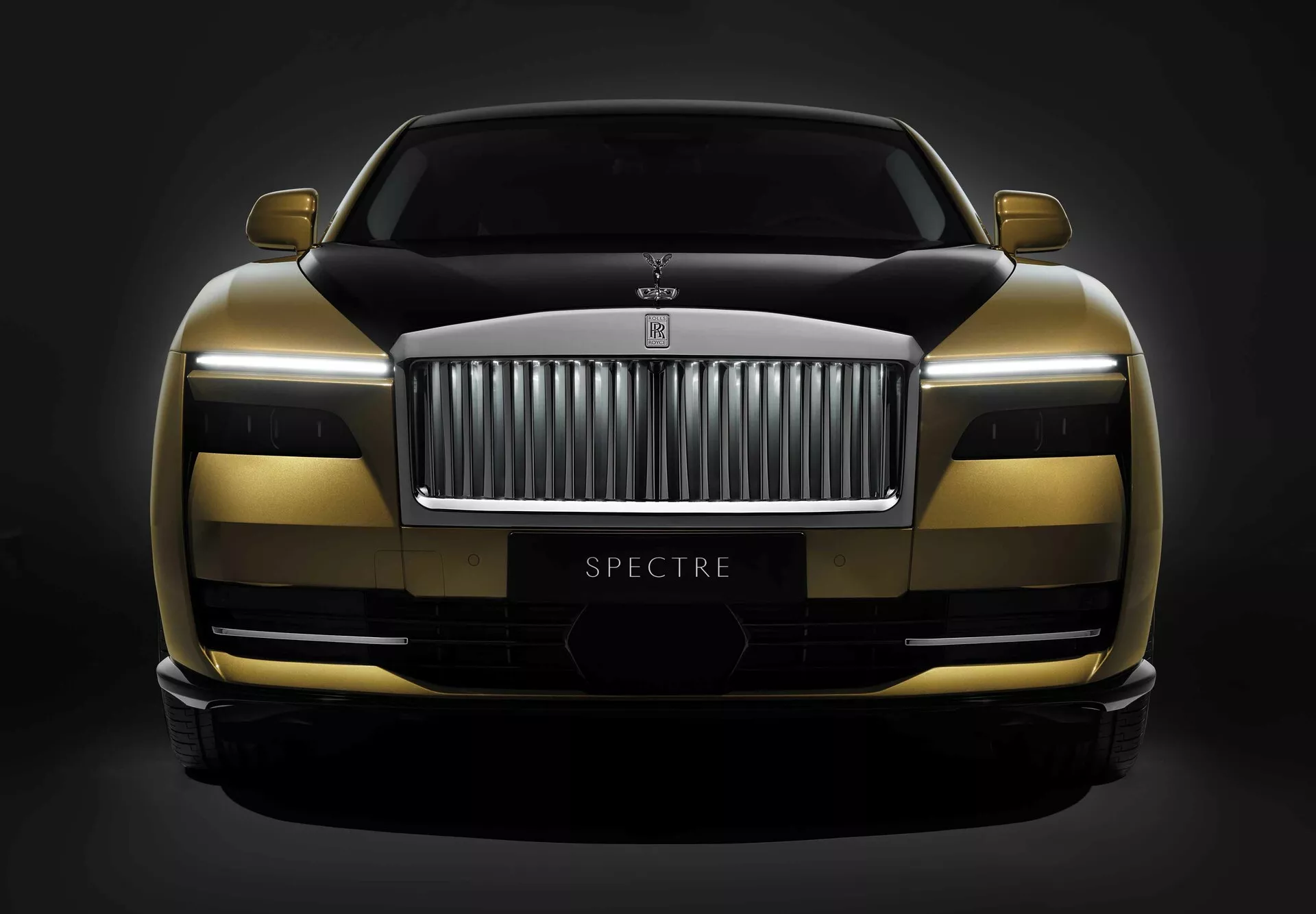 Rolls-Royce Spectre 1. generacije 2023 prikazuje dvobarvno shemo barv, napredno tehnologijo LED žarometov in aerodinamično obliko z vidnim imenom modela 'SPECTRE', posneto iz nizkega sprednjega kota na temnem ozadju.