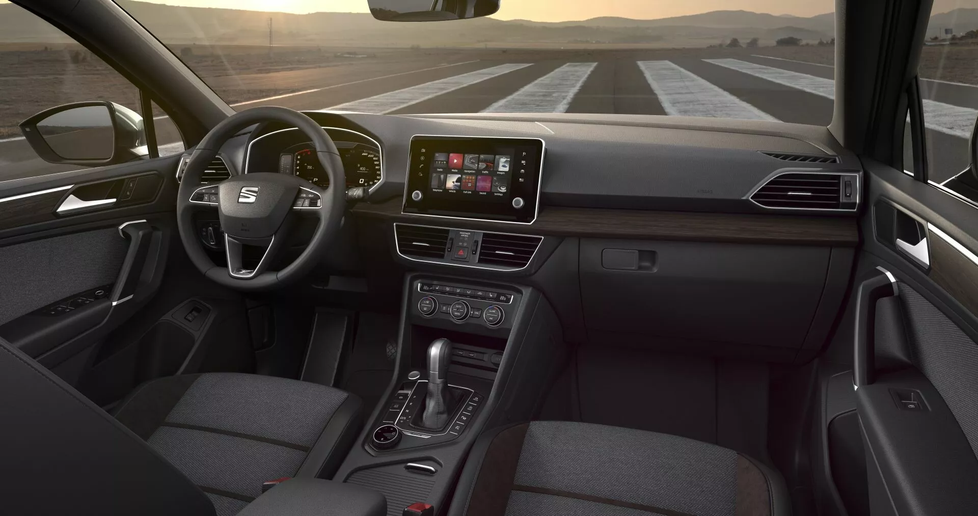 Notranjost Seat Tarraco 2019 z vidika zadnjega sedeža, prikazuje večfunkcijski volan, centralni zaslon na dotik za infotainment, gumbe za nadzor klime, ročico avtomatskega menjalnika in sedež potnika oblazinjen s tkanino, pod diskretno notranjo osvetlitvijo
