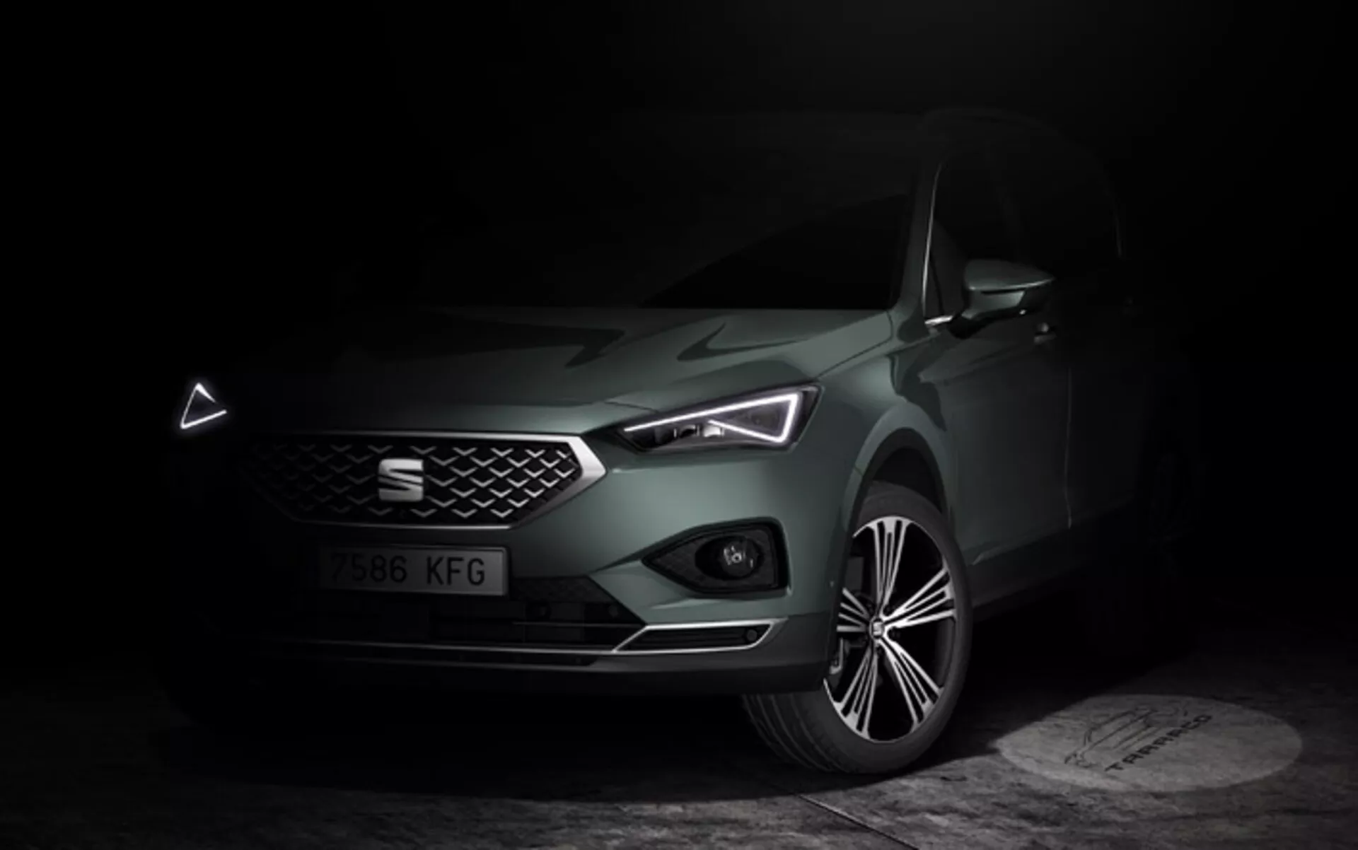 Seat Tarraco 2019 prikazan v temnem, razpoloženem okolju, ki poudarja njegov eleganten sprednji del, geometrijsko rešetko, LED žaromete z značilnimi dnevnimi lučmi in kiparsko oblikovano pokrovom motorja, skupaj z detajliranim prikazom lita platišča, ki poudarja luksuzne in sodobne tehnološke značilnosti avtomobila
