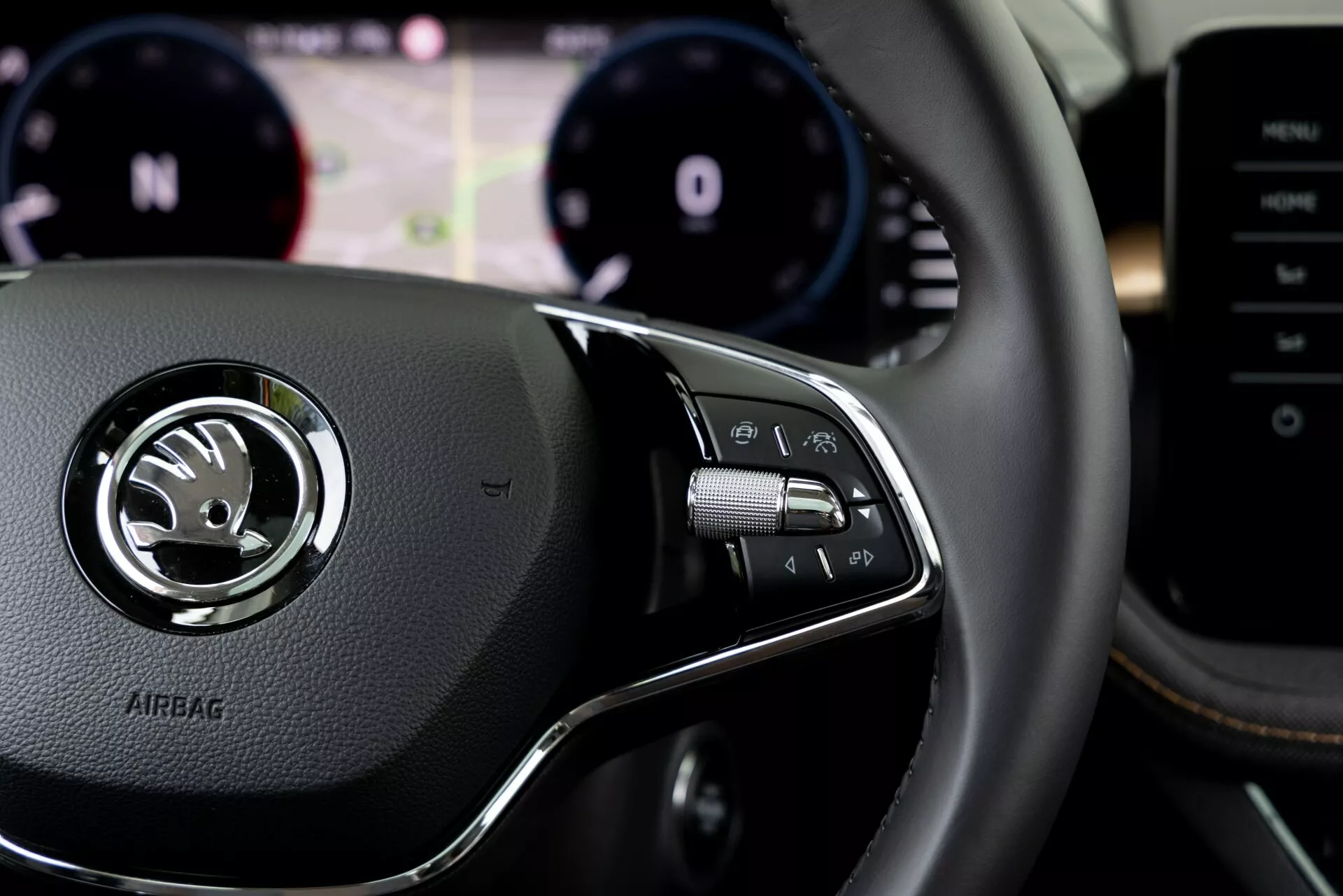 Podrobnejši pogled na notranjost Škode Fabia 2021 iz voznikove perspektive, s poudarkom na desnih kontrolah na volanu. Usnjen volan ima sredinsko srebrno značko z logotipom Škoda in vtisnjen napis 'AIRBAG' spodaj. Na desnem kontrolnem sklopu so gumbi za multimedijski sistem, prostoročne telefonske klice ali glasovne ukaze, z vidnimi vzvodi za ležaje za volanom. Zabrisano ozadje prikazuje nadzorno ploščo z merilnikom vrtljajev in merilnikom hitrosti ter osrednjim digitalnim zaslonom za pomoč pri vožnji. Delno vidna je tudi sredinska konzola ali multimedijski sistem na desni strani.