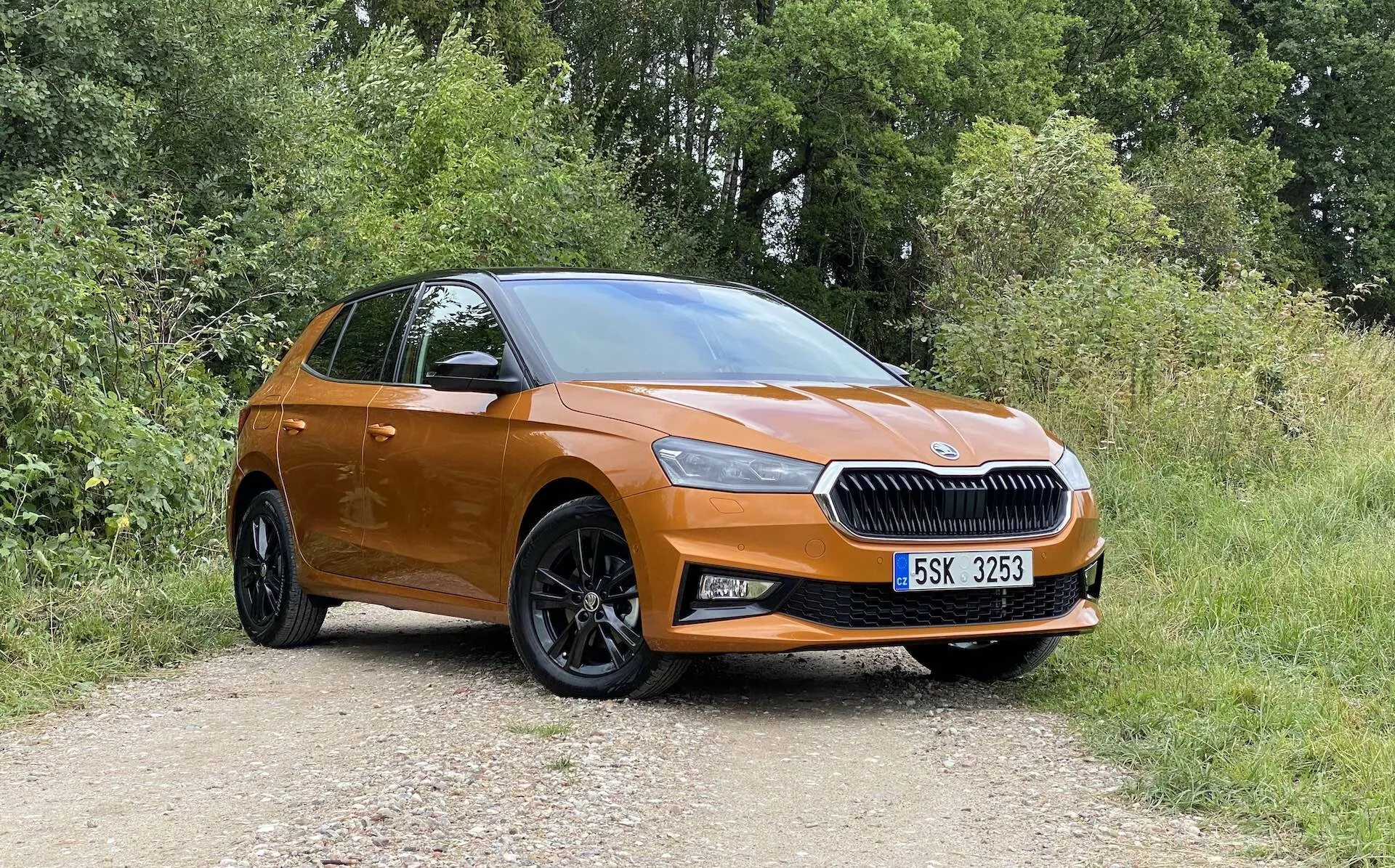 Popoln bočni profil modernega, oranžno obarvanega Škoda Fabia 2021 hatchbacka, parkiranega na neasfaltirani poti z vegetacijo v ozadju, posnet pod rahlim poševnim kotom spredaj, poudarjajoč voznikovo stran. Značilnosti vključujejo izrazito masko z osrednjim logotipom blagovne znamke, elegantne žaromete, meglenke, velika črna večkraka platišča iz lahke litine, barvno usklajene kljuke vrat in ogledala, rahlo padajočo linijo strehe ter namig na zadnja vrata in svetlobni sklop.