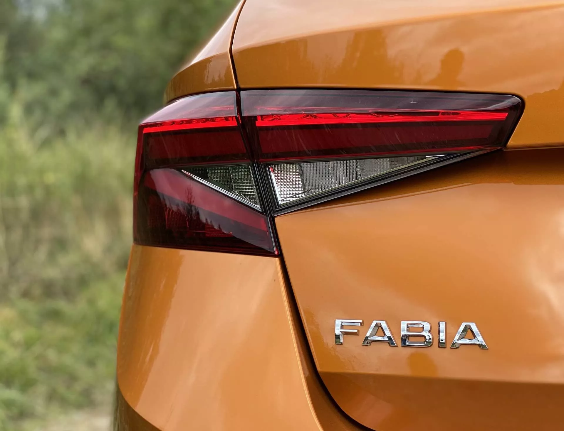 Posnetek zadnjega kota Škode Fabia 2021 od blizu, ki poudarja ostro, kotno zasnovo zadnje LED luči in značilno blagovno znamko 'FABIA' na sijočem, kovinsko oranžnem telesu. Gladke krivine in nežni izboklini okoli zadnje luči prispevajo k sodobni estetiki, zajeti iz rahlo dvignjenega kota z majhno globino polja.